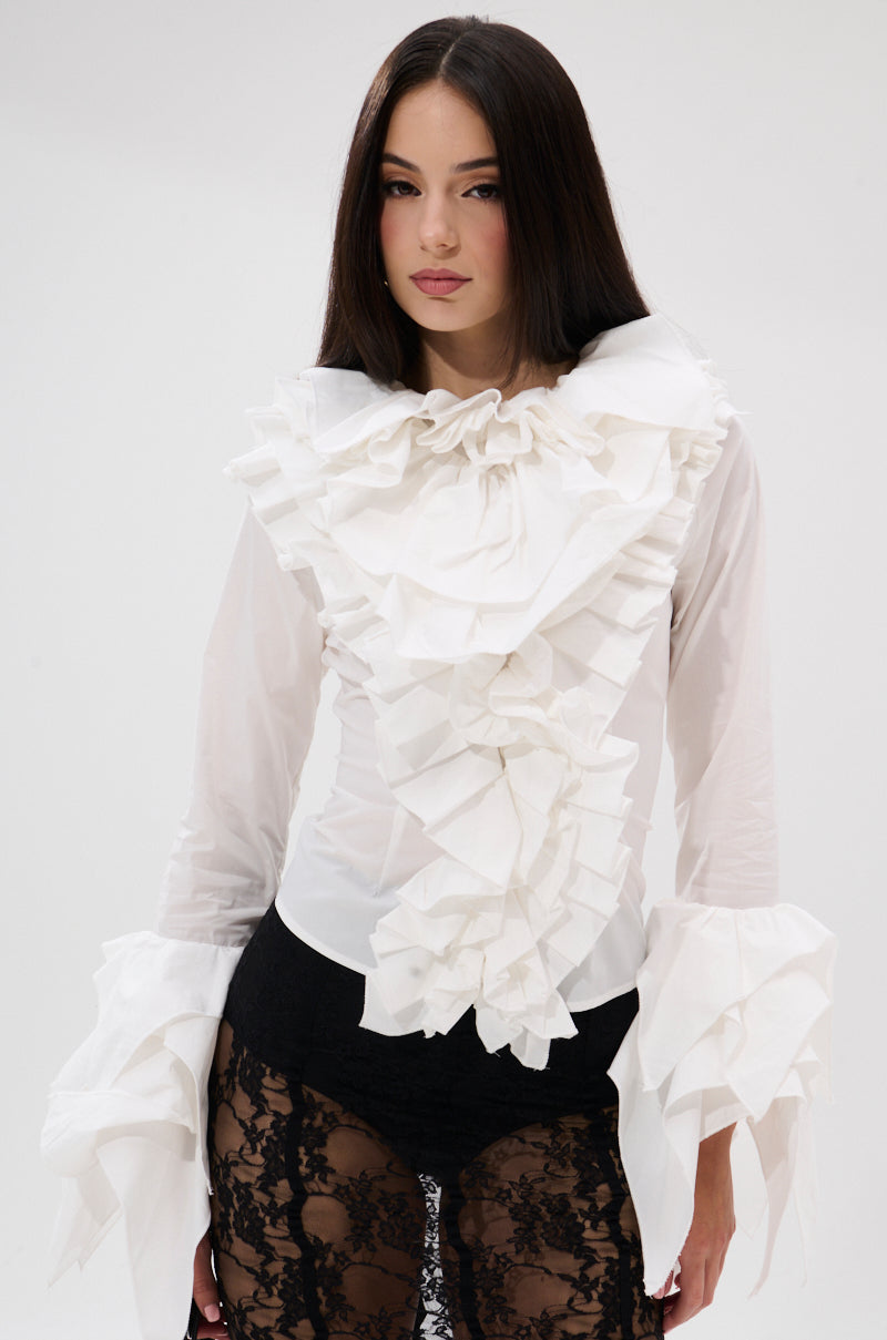 A woman with long dark hair wears the OVER THE TOP RUFFLE BLOUSE, a white top with dramatic sleeves and oversized frills, styled with black lace pants against a plain, light-colored background. 1