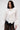 A woman with long dark hair wears the OVER THE TOP RUFFLE BLOUSE, a white top with dramatic sleeves and oversized frills, styled with black lace pants against a plain, light-colored background.