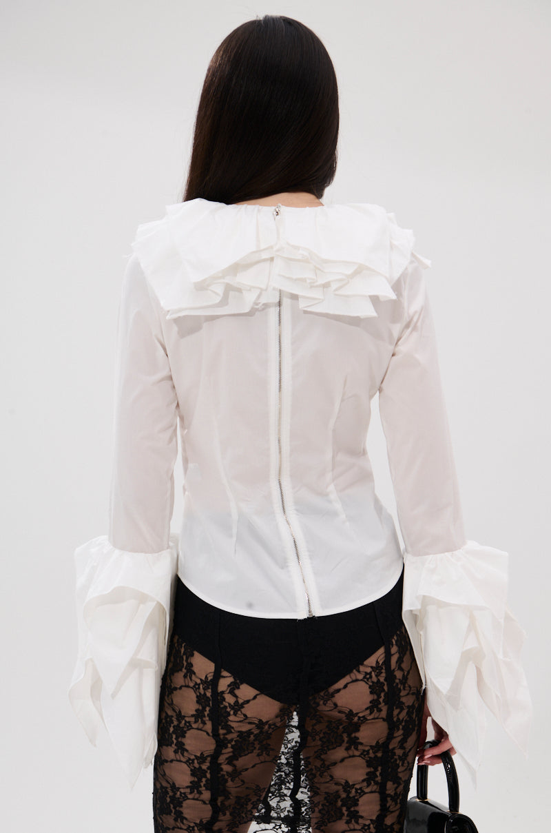 A woman with long dark hair, seen from behind, wears the OVER THE TOP RUFFLE BLOUSE with ruffled cuffs and collar, paired with a sheer black lace skirt and holding a black handbag against a plain light background. 3