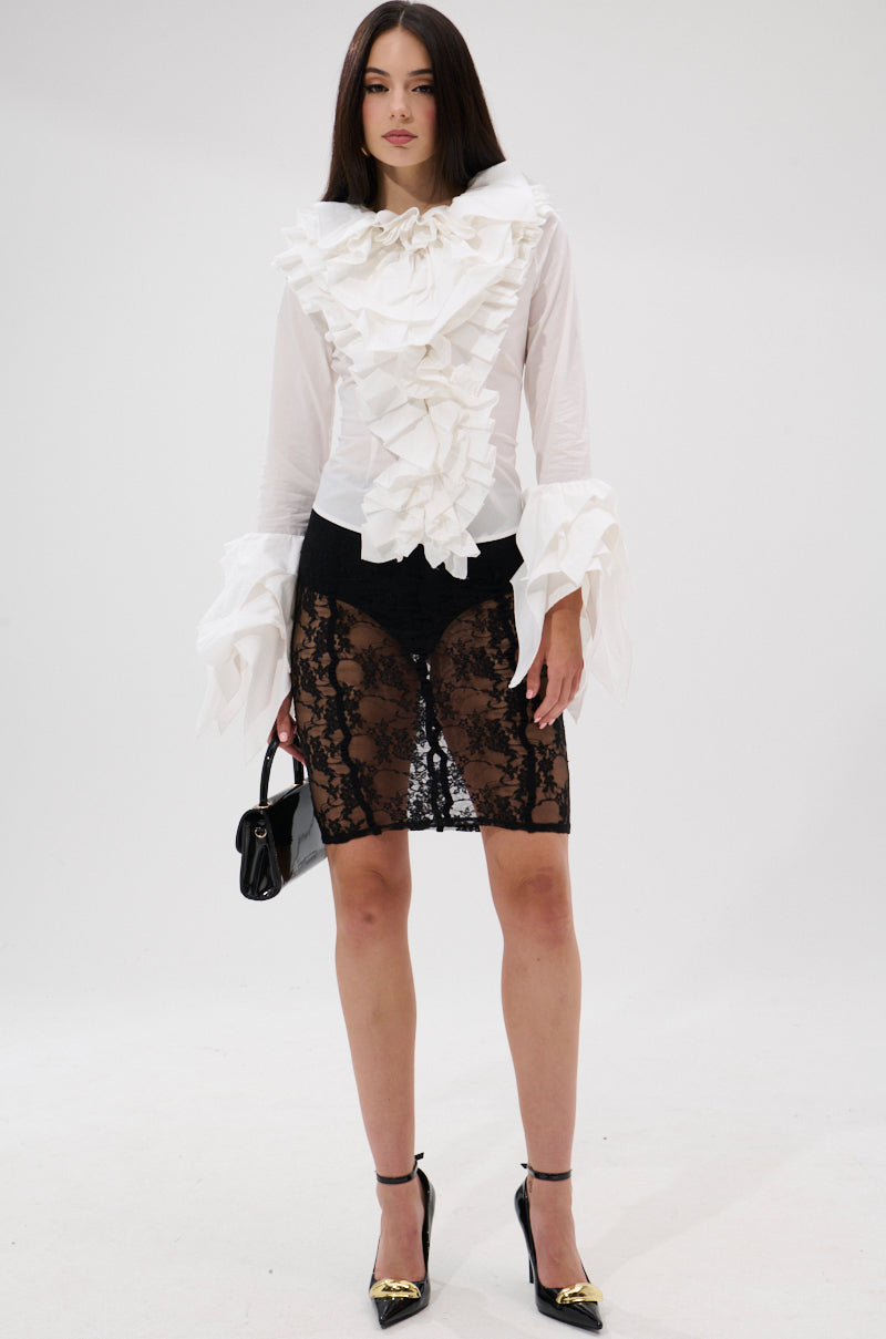 A woman poses before a white background in the AKIRA OVER THE TOP RUFFLE BLOUSE, paired with a black lace skirt overlay, black heels with gold accents, and carrying a small black handbag. 5