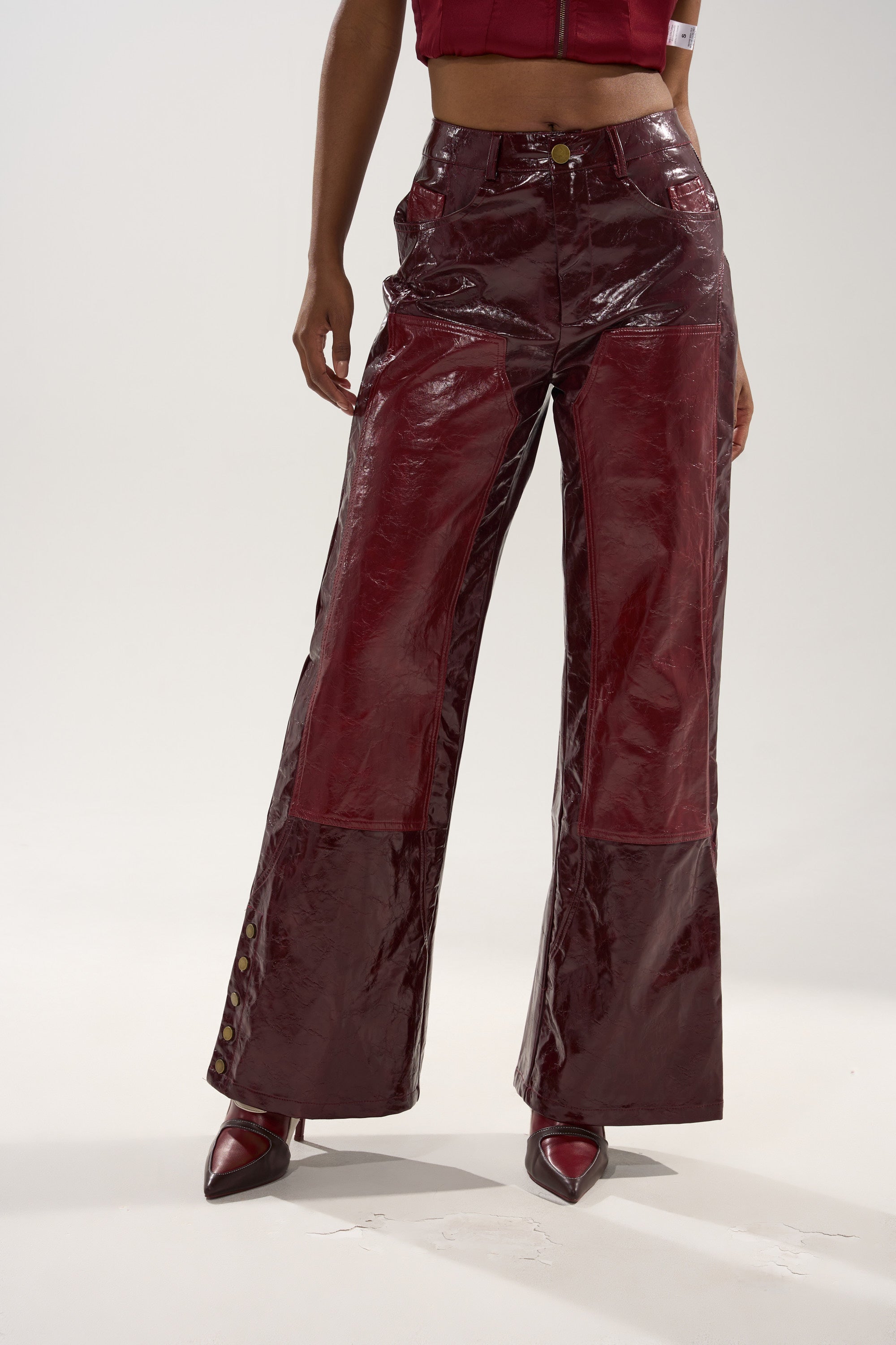 A person stands against a plain background wearing the MIXED EMOTIONS FAUX LEATHER TROUSER—a shiny, burgundy, high-rise, straight-leg pant—paired with pointed-toe heels and a sleeveless crop top. Their left hand rests confidently on their hip.