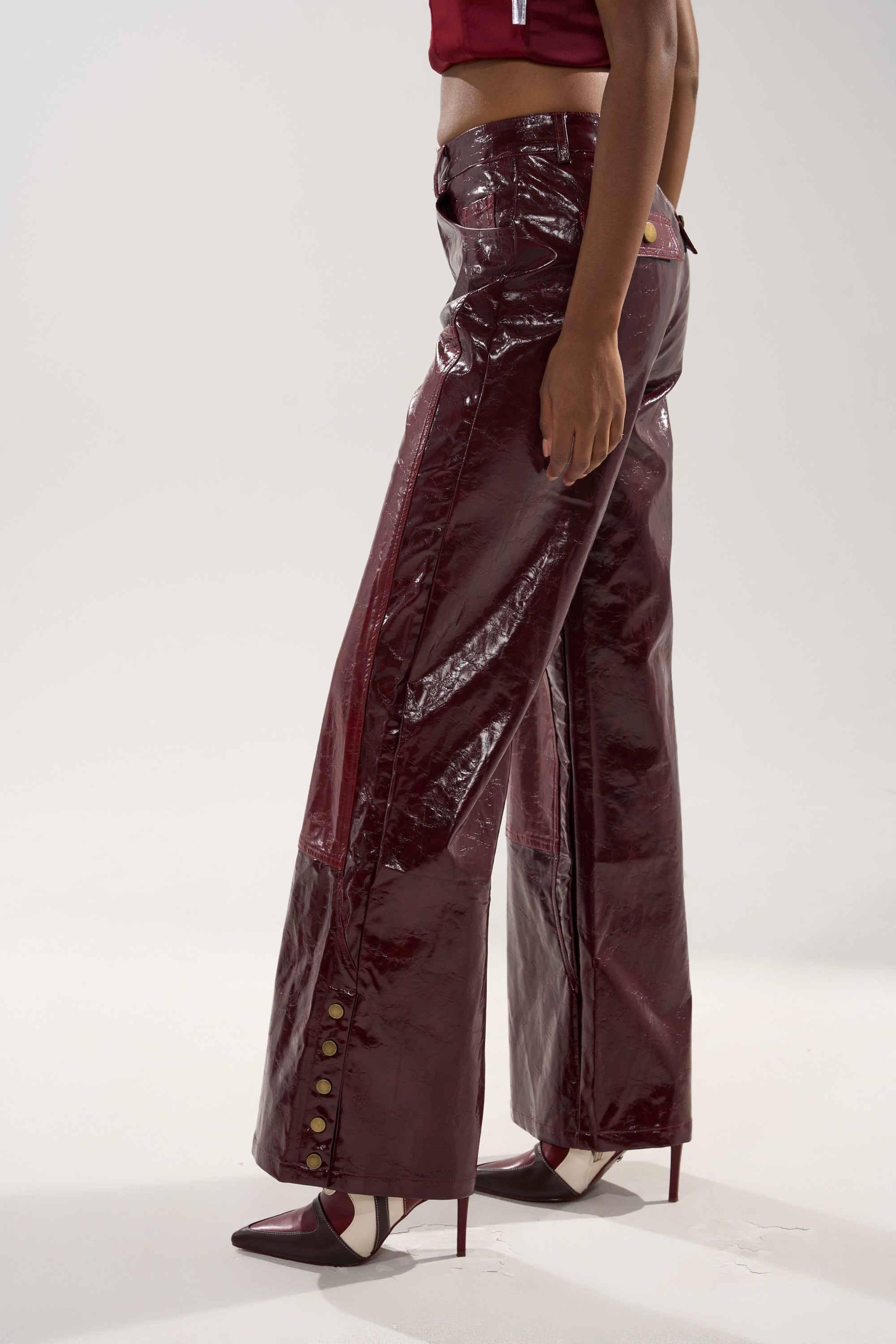 Wearing the MIXED EMOTIONS FAUX LEATHER TROUSER—shiny burgundy high-rise, straight-leg with ankle buttons—paired with pointed heels, a person stands against a plain light background, shown from chest down.
