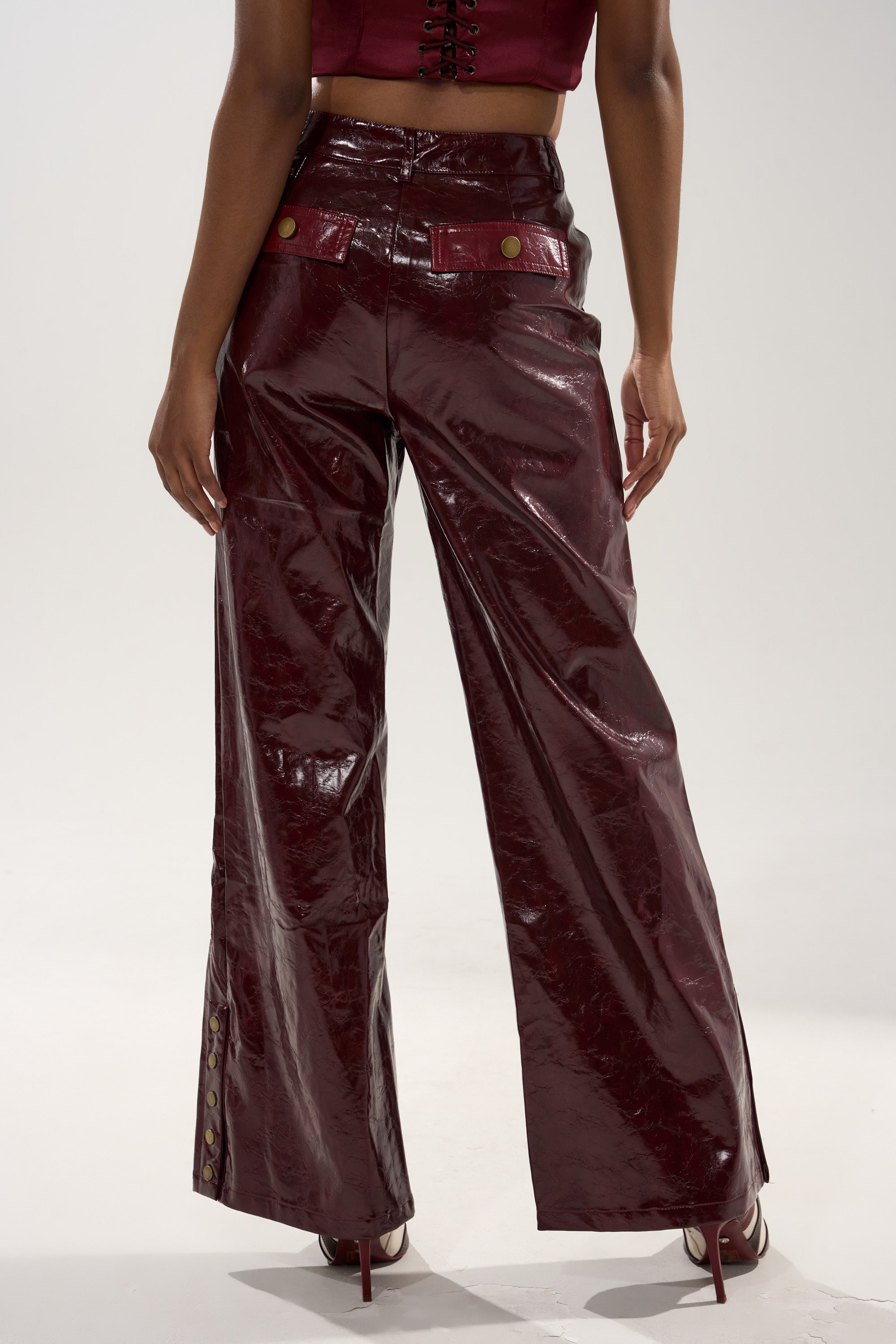 A person stands facing away in the MIXED EMOTIONS FAUX LEATHER TROUSER—shiny, dark red, high-rise with gold buttons and a wide-leg silhouette—paired with a matching cropped top and heels against a plain light background.