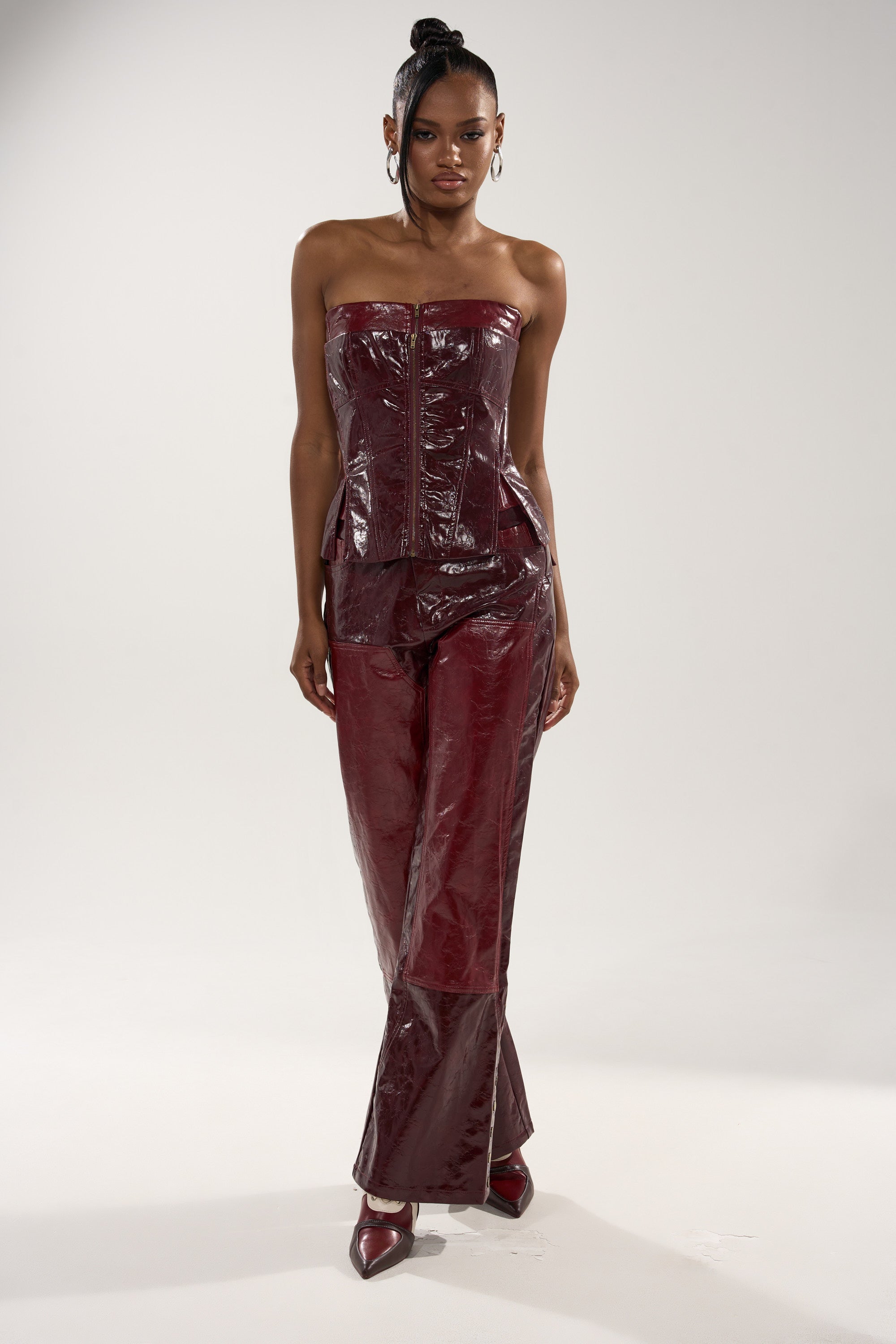 A woman poses against a plain background in the MIXED EMOTIONS FAUX LEATHER TROUSER, styled with a shiny, burgundy strapless corset, pointed heels, hoop earrings, and her hair in a high ponytail.