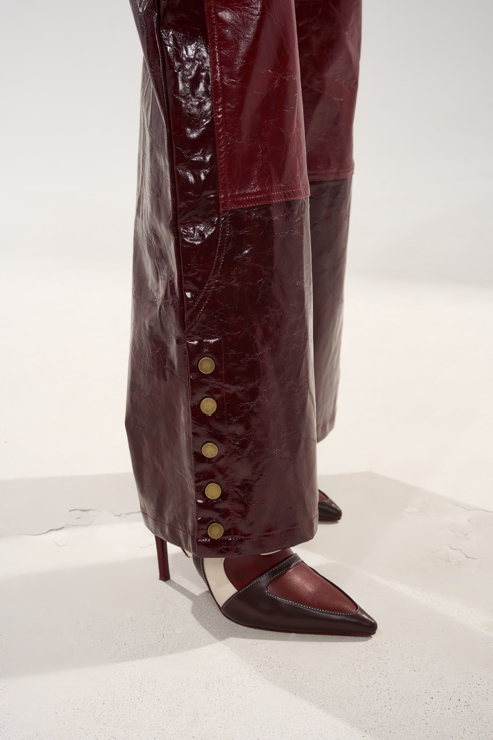 A close-up of someone wearing the MIXED EMOTIONS FAUX LEATHER TROUSER in glossy dark red with gold snap buttons along the sides and pointed red-and-black heels. The straight-leg silhouette pops against a plain, light background.