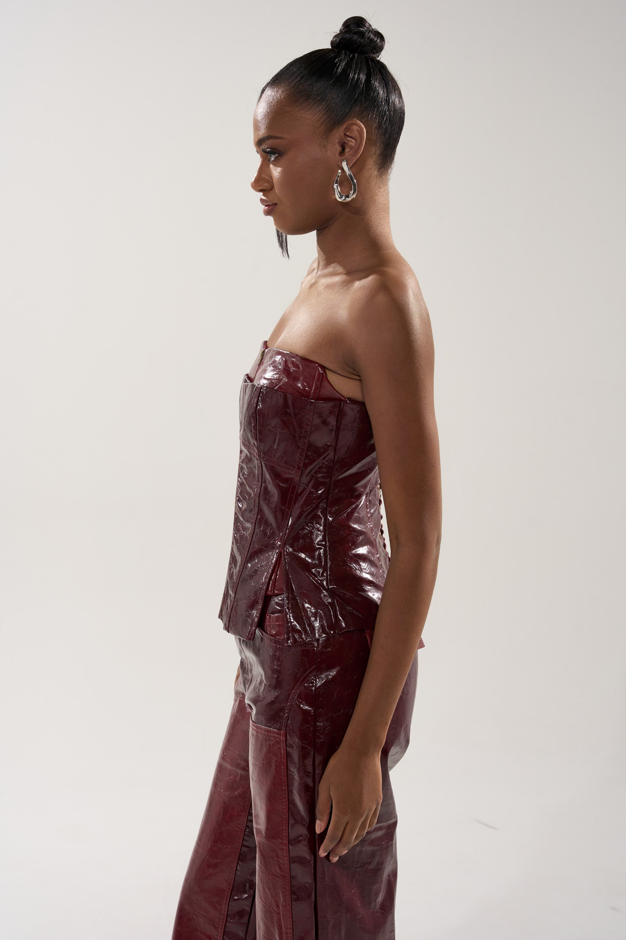 A woman with a sleek bun wears the MIXED EMOTIONS FAUX LEATHER CORSET, a shiny burgundy strapless top, paired with matching pants. She stands in profile against a light background, accessorized with large hoop earrings.