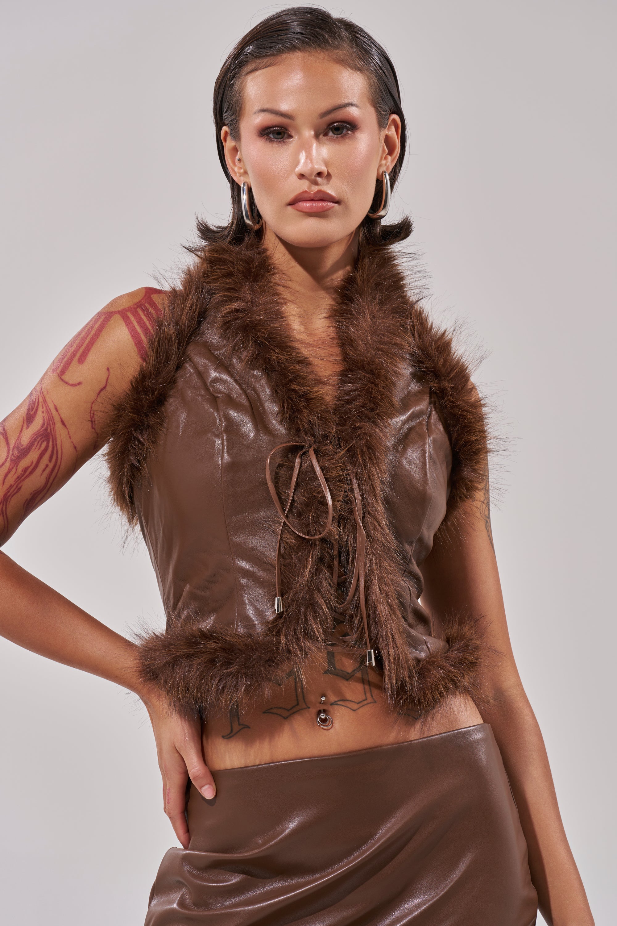 A woman with slicked-back hair wears the CANT MISS ME FAUX LEATHER FUR CORSET and matching skirt, showing her arm tattoo, hoop earrings, and navel piercing as she stands against a plain light gray background. 0