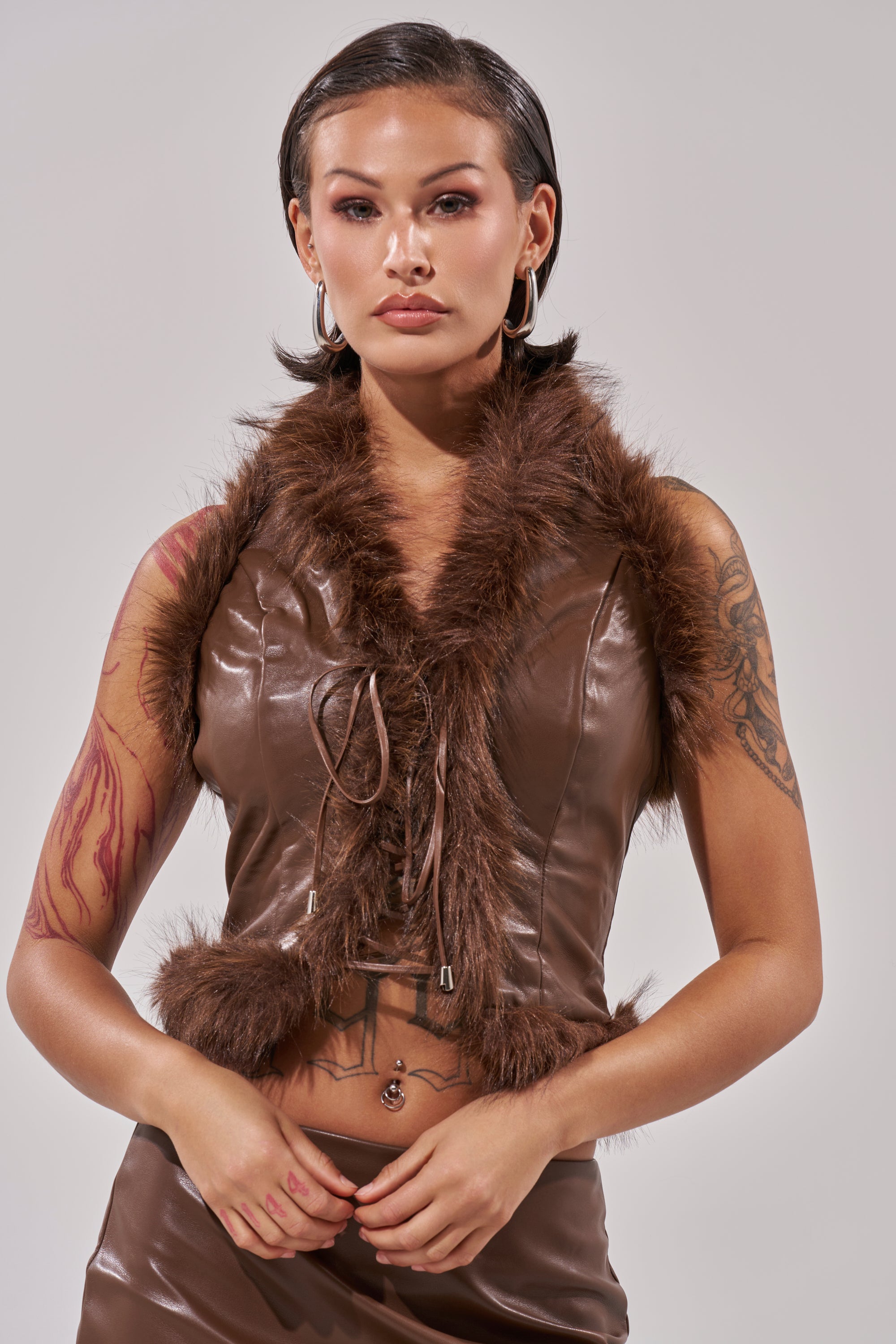 A woman with short dark hair wears the CANT MISS ME FAUX LEATHER FUR CORSET and a matching skirt, showing tattoos, hoop earrings, and a belly button piercing, posed against a light background. 2