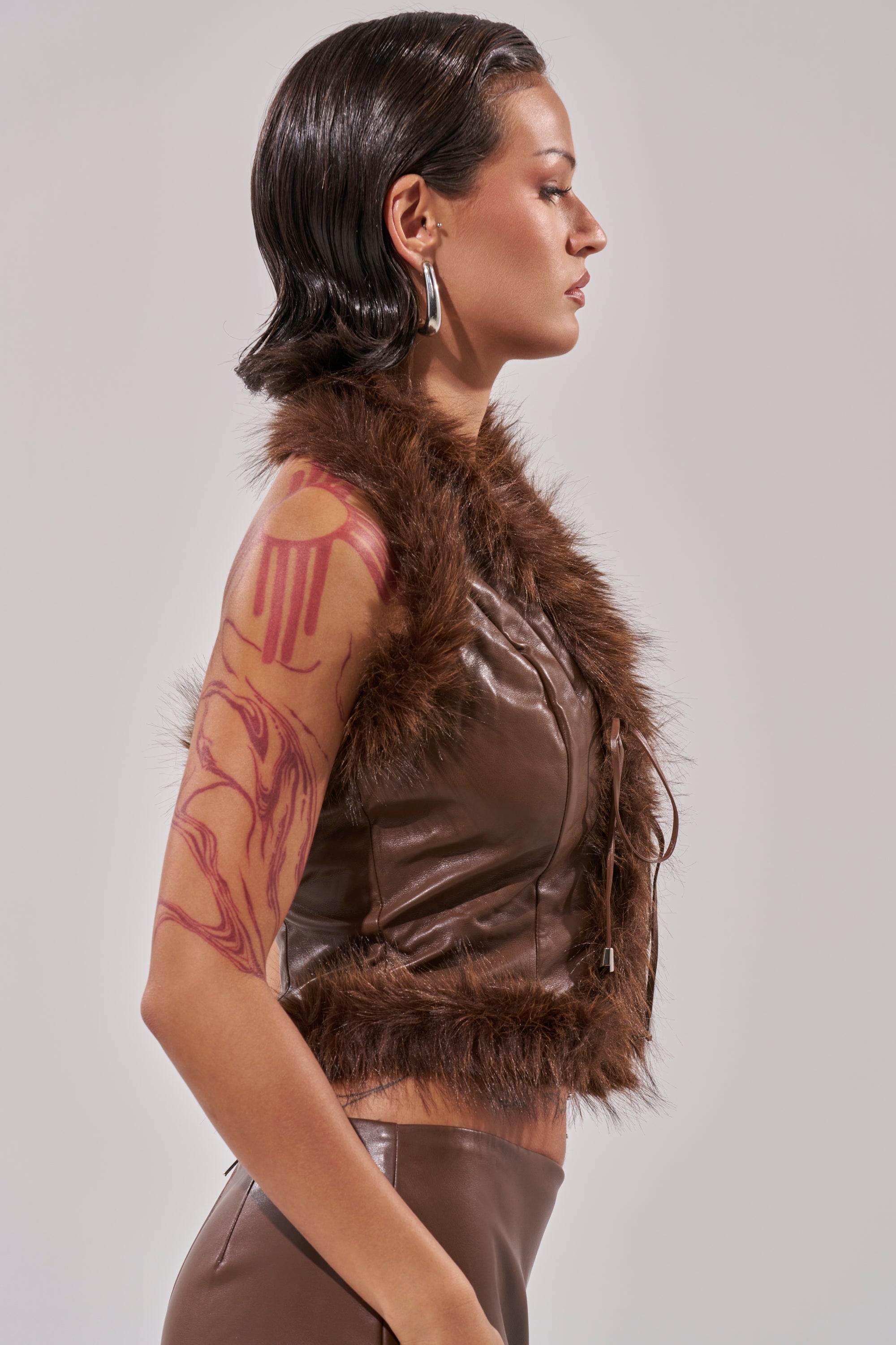 A woman with slicked-back dark hair and hoop earrings poses in profile, showcasing the CANT MISS ME FAUX LEATHER FUR CORSET and matching pants. A bold red abstract tattoo is visible on her upper arm. 3