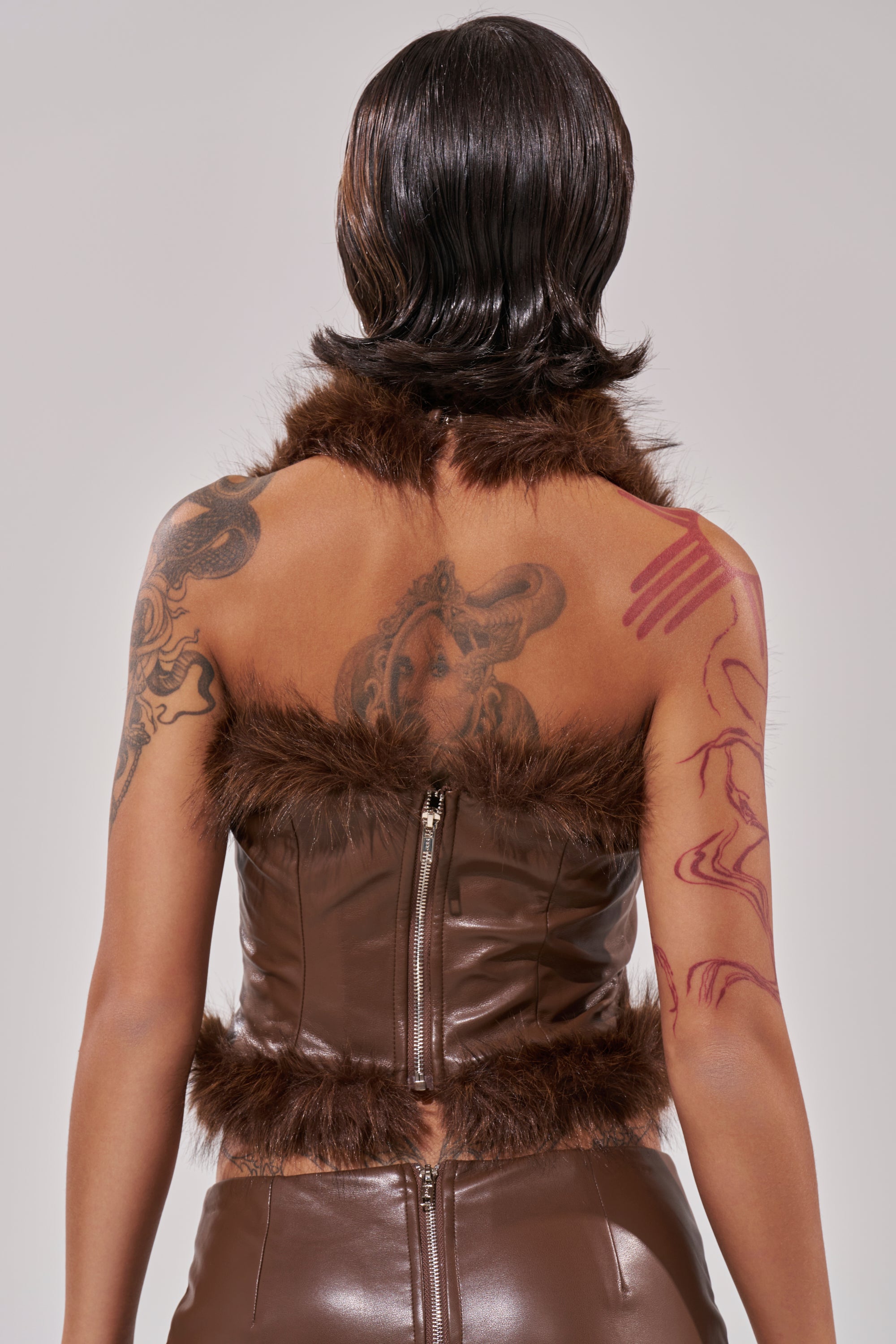 A person with short, slicked-back hair is seen from behind wearing the CANT MISS ME FAUX LEATHER FUR CORSET, featuring brown faux leather with fur trim and a zipper, highlighting their tattooed back and arms. 4