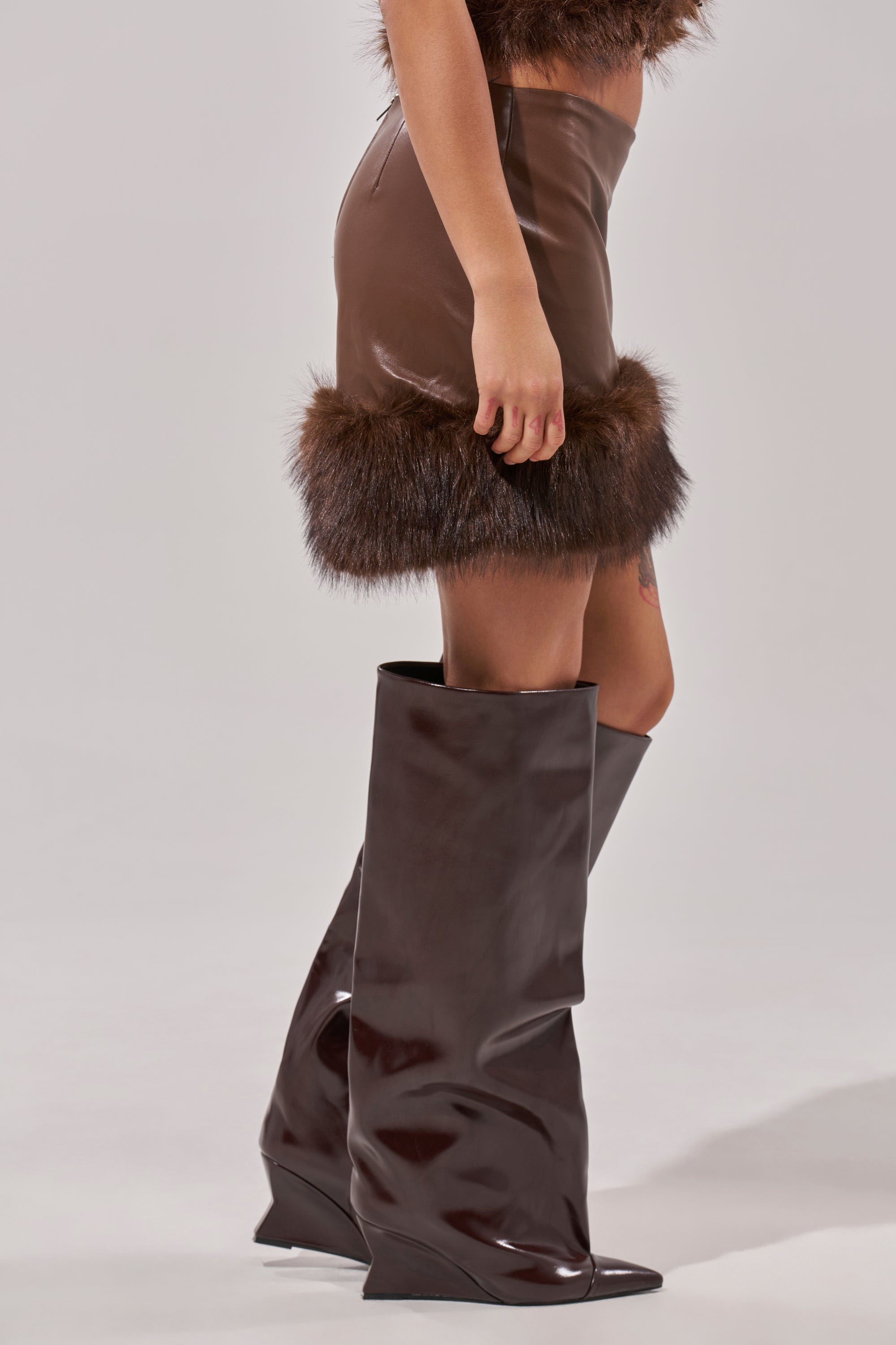 Wearing the CANT MISS ME FAUX LEATHER MINI SKIRT with fur-trimmed hem and shiny, oversized dark brown wedge boots. The background is plain and light-colored. 2