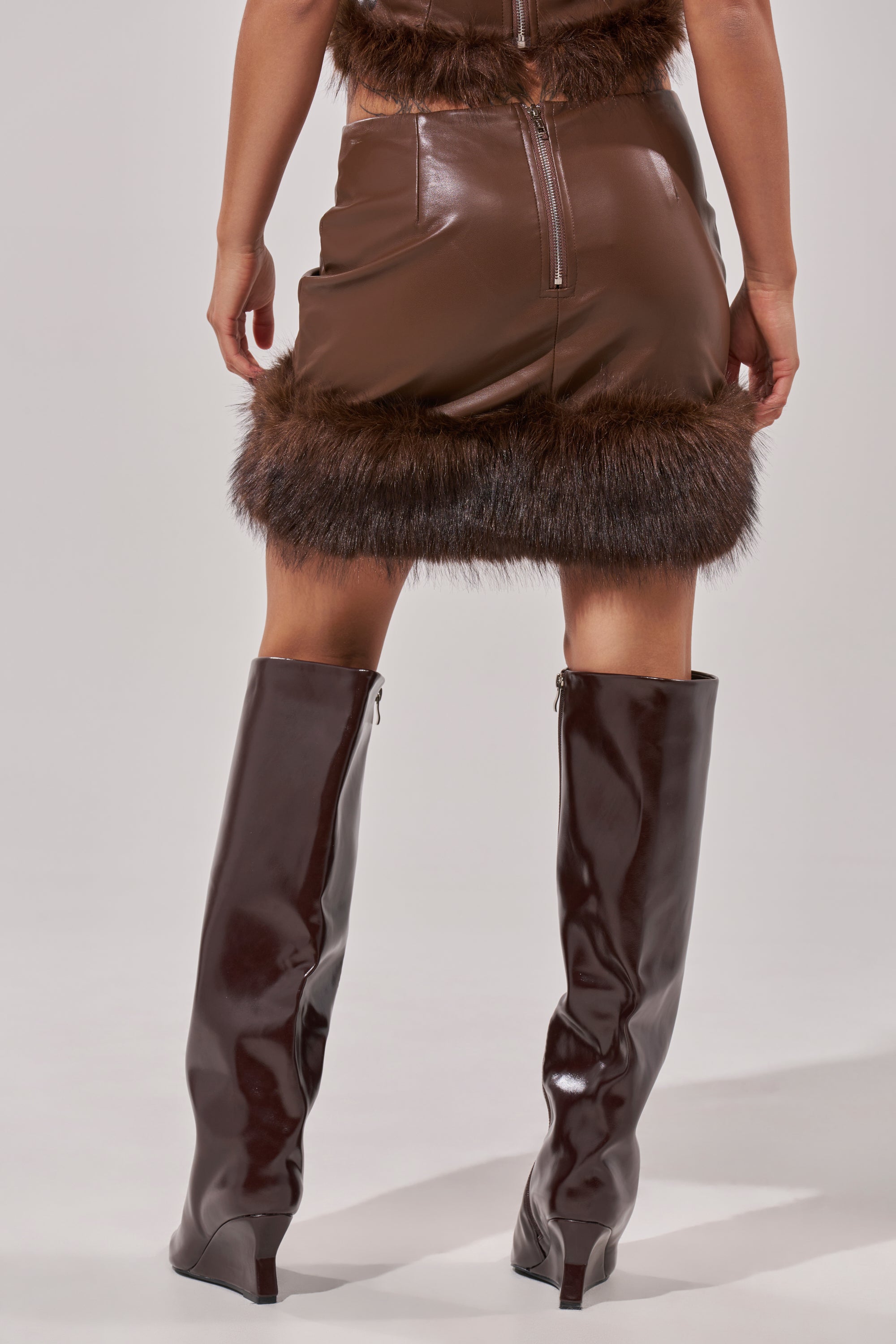 A person wearing the CANT MISS ME FAUX LEATHER MINI SKIRT with fur trim, paired with tall shiny brown boots, is shown from the back against a neutral background. 3