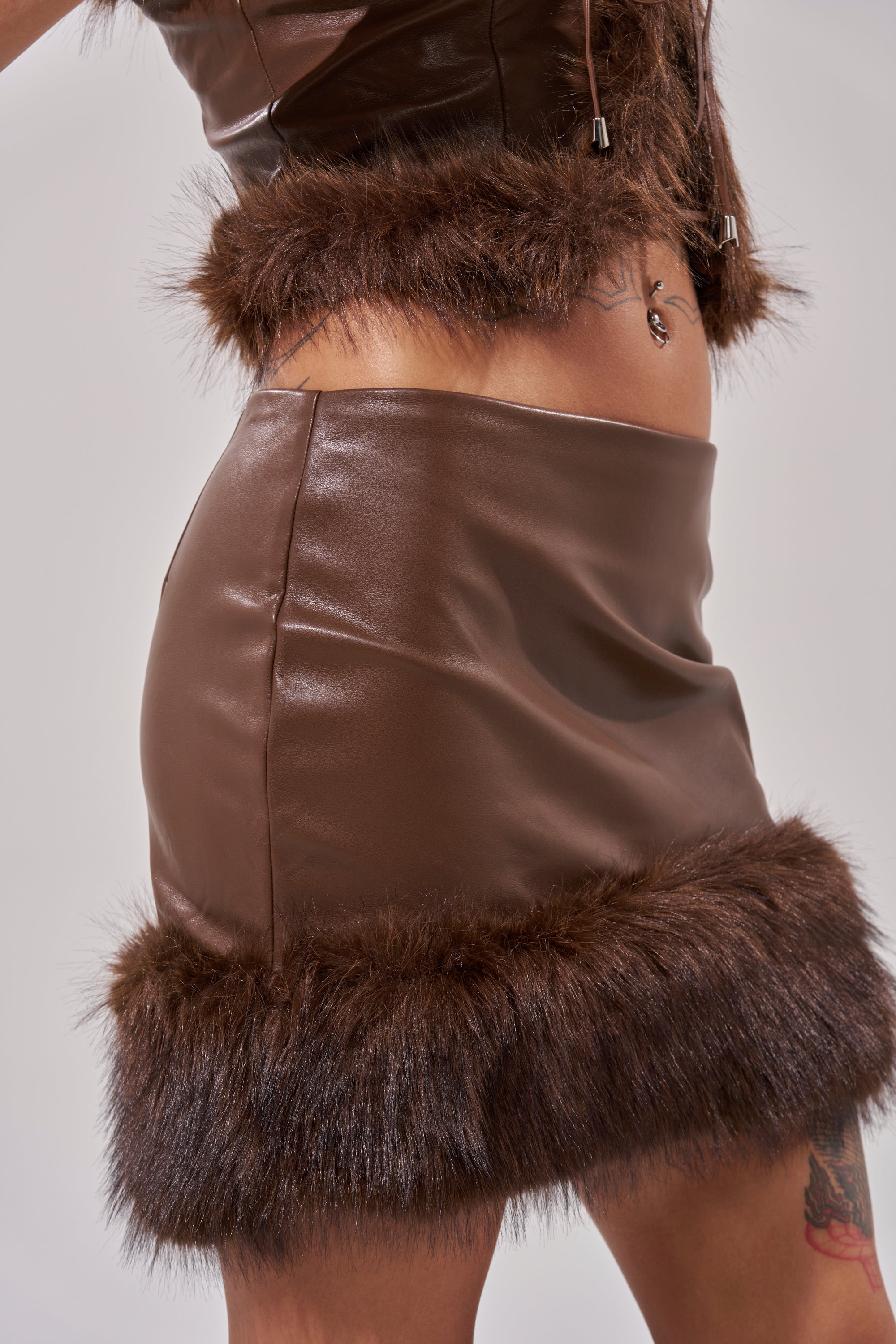 Wearing the CANT MISS ME FAUX LEATHER MINI SKIRT with a matching brown faux fur-trimmed top, the cropped image highlights the skirt’s luxurious texture and a thigh tattoo, focusing on the torso and upper legs. 6