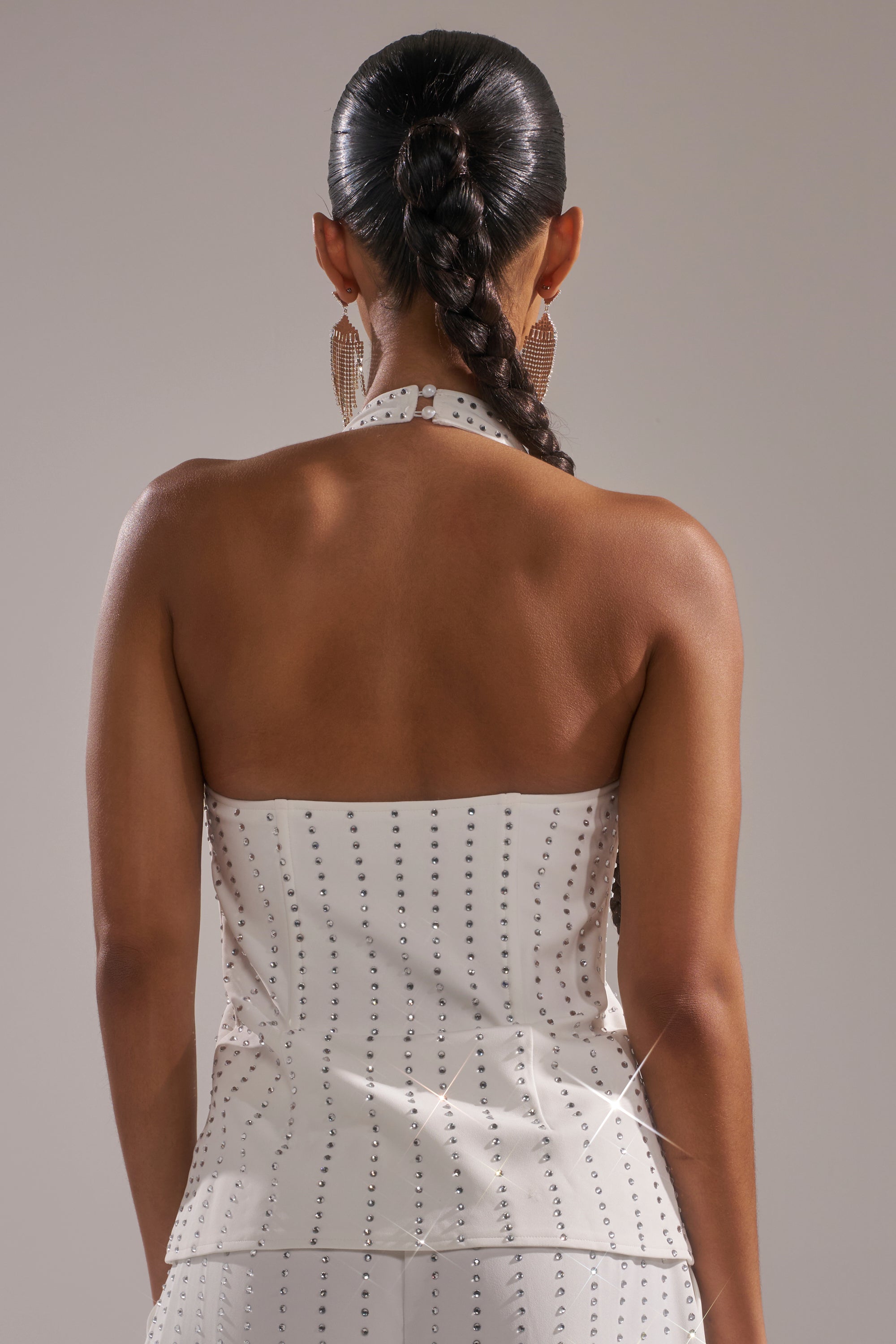 A woman with a sleek braided ponytail is seen from the back, wearing the NEW YEARS QUEEN VEST IN WHITE—a strapless top with a subtle dotted pattern and tailored fit—paired with large textured earrings against a neutral background. 3