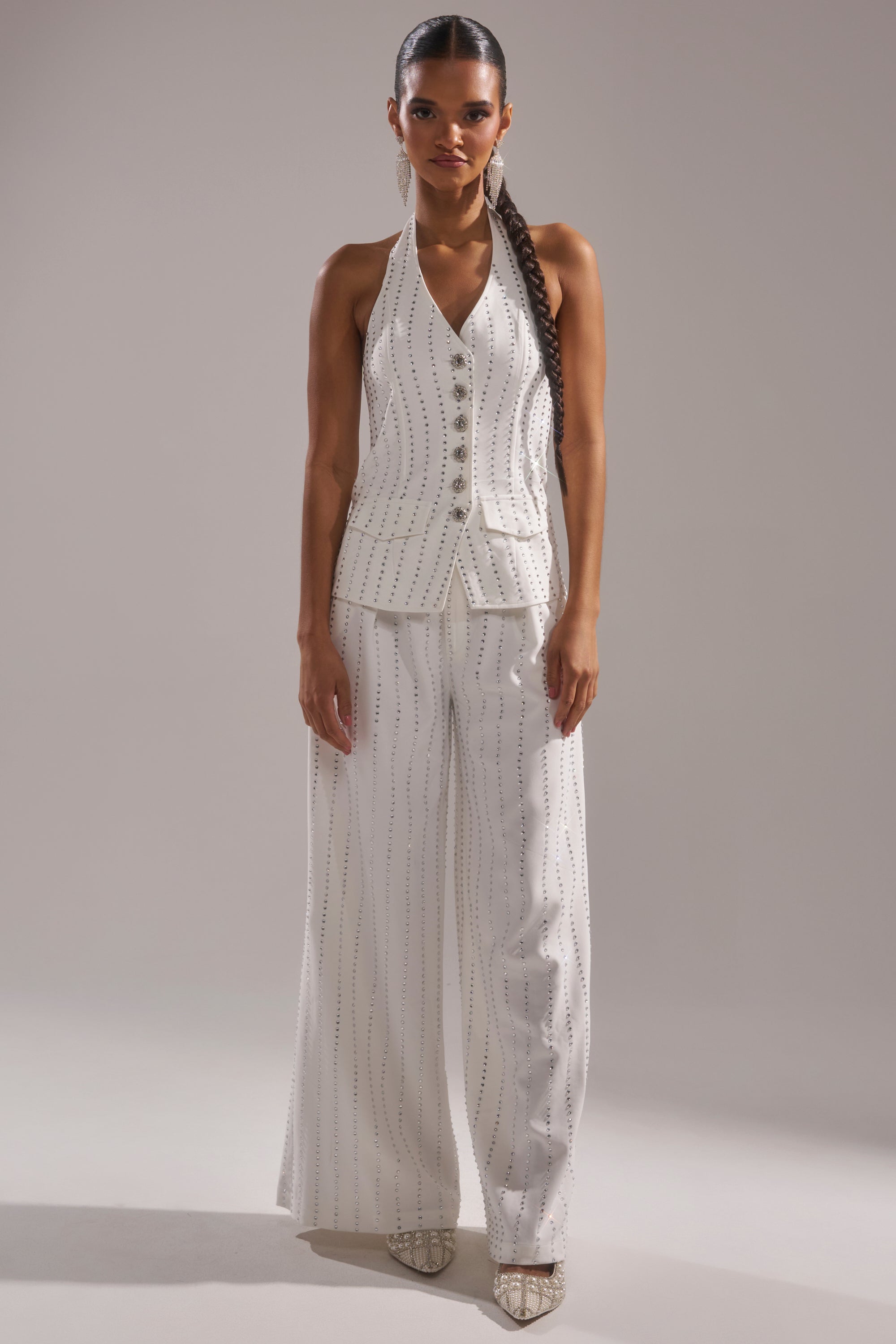 A woman wears the NEW YEARS QUEEN VEST IN WHITE, a sleeveless white rhinestone vest with silver buttons and vertical dotted lines, paired with matching wide-leg pants, pointed-toe heels, and her hair in a long braid. 5