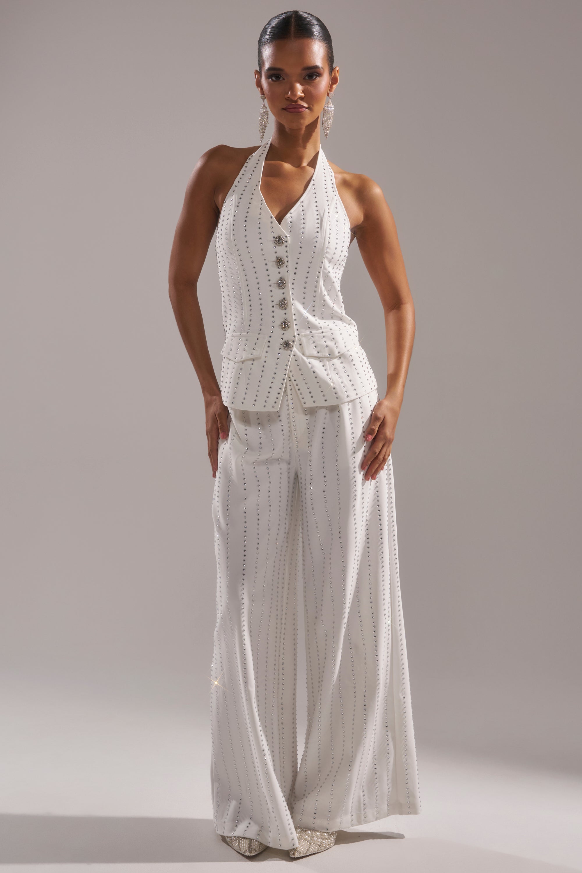 A person stands confidently in a sleeveless white halter top and NEW YEARS QUEEN TROUSER—wide-leg pants featuring vertical silver-studded lines—against a plain light gray background. 0