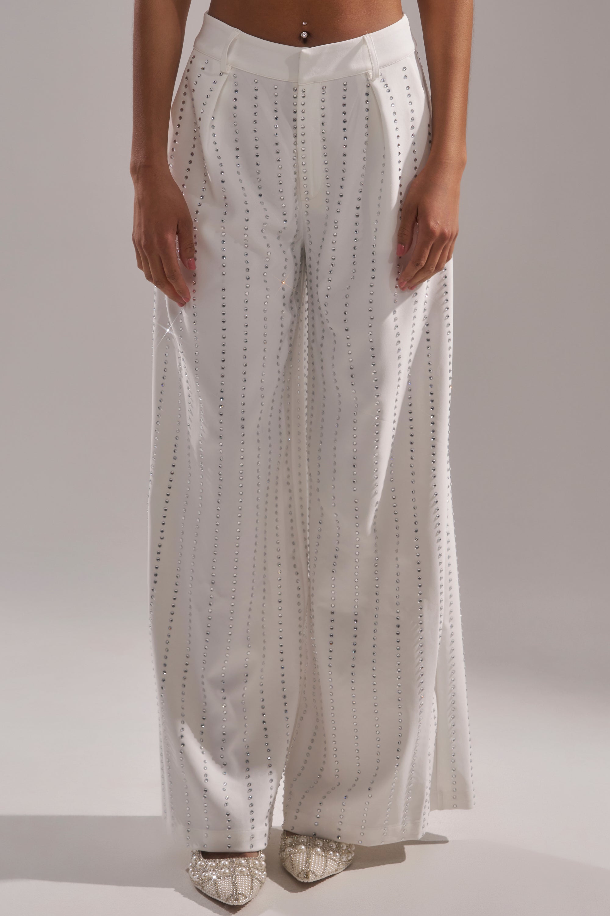 Wearing the NEW YEARS QUEEN TROUSER—high-rise, wide-leg white pants with vertical sequin details—and pointed-toe snakeskin shoes, she stands out against a plain background. 1