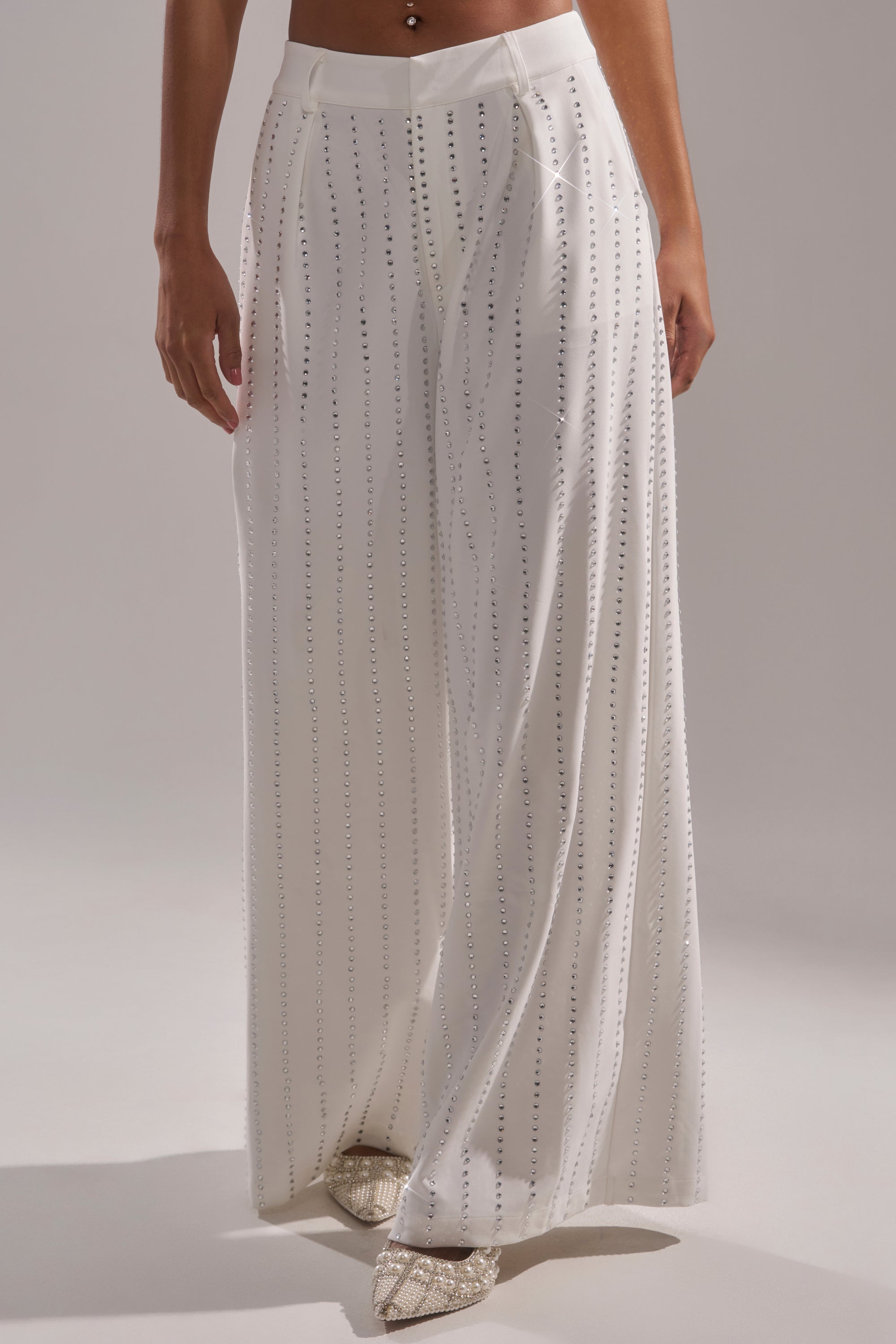 A person wears the NEW YEARS QUEEN TROUSER—high-rise, wide-leg pants with vertical silver embellishments—paired with sparkly pointed-toe shoes. A softly lit, neutral background highlights this elegant look. 2