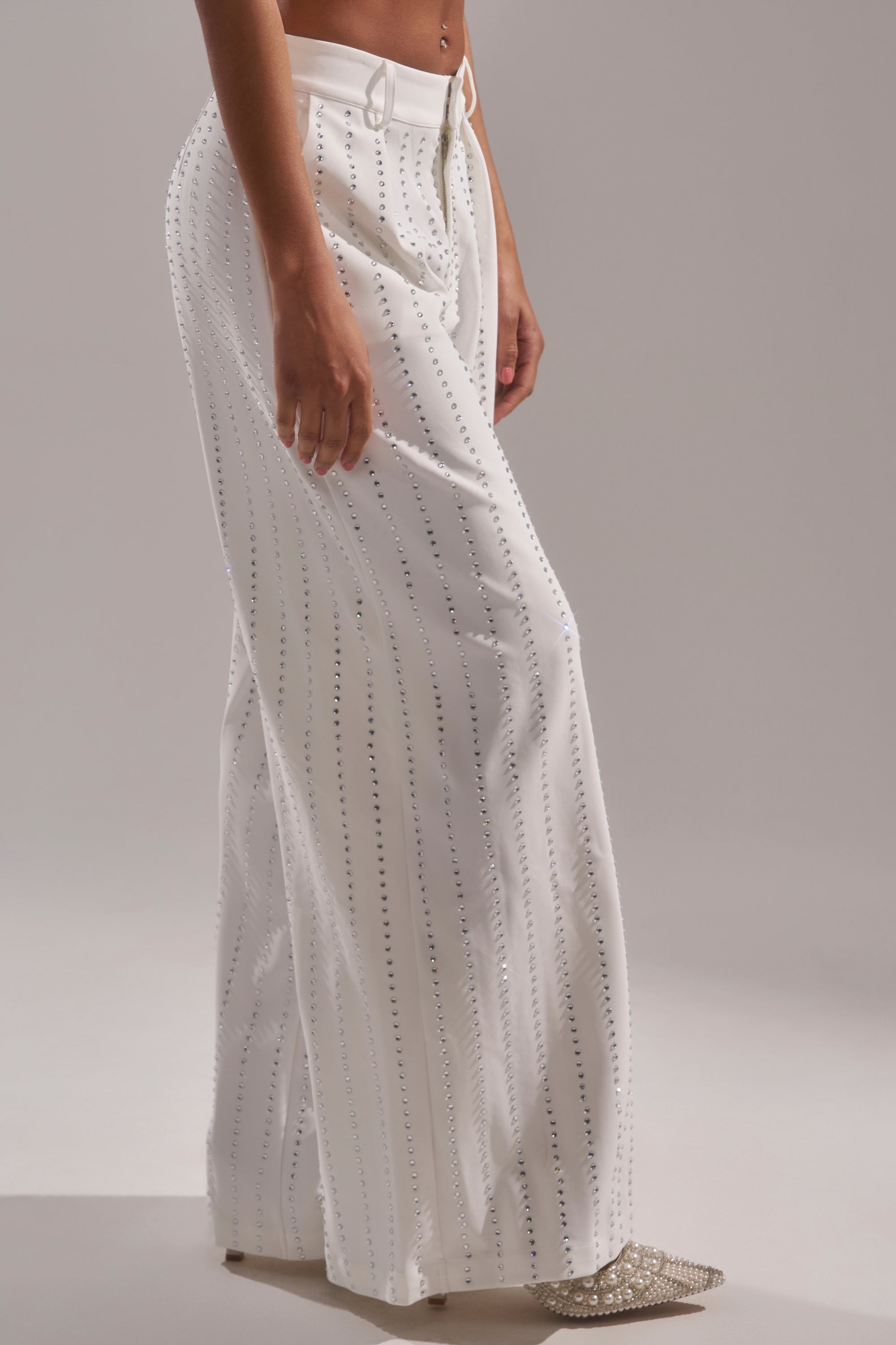 Wearing the NEW YEARS QUEEN TROUSER—high-rise, wide-leg rhinestone trousers with vertical studs—and pointed embellished shoes, a person stands against a plain light background. 3