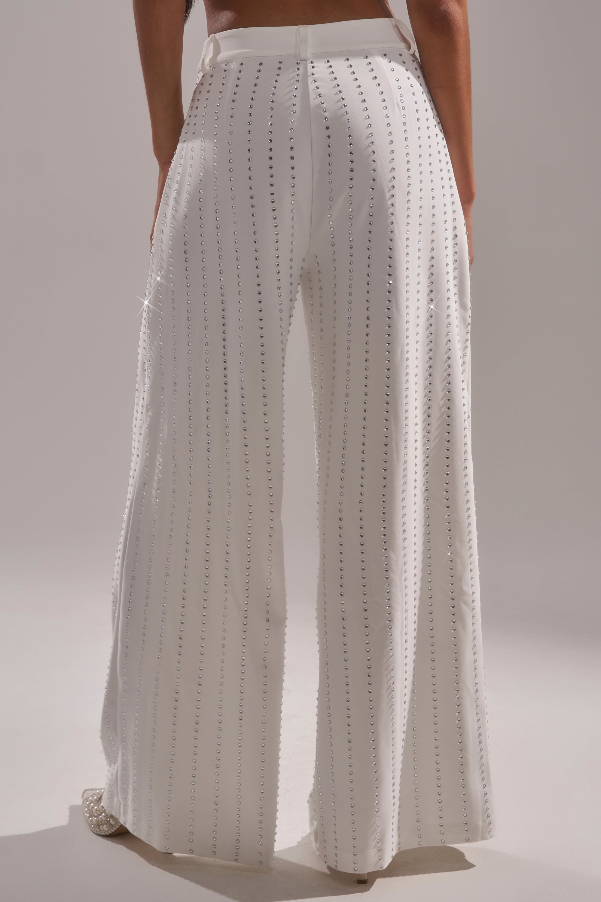 Wearing the NEW YEARS QUEEN TROUSER—high-waisted, wide-leg white pants with vertical rhinestone rows—a person stands with their back to a plain light background. 4