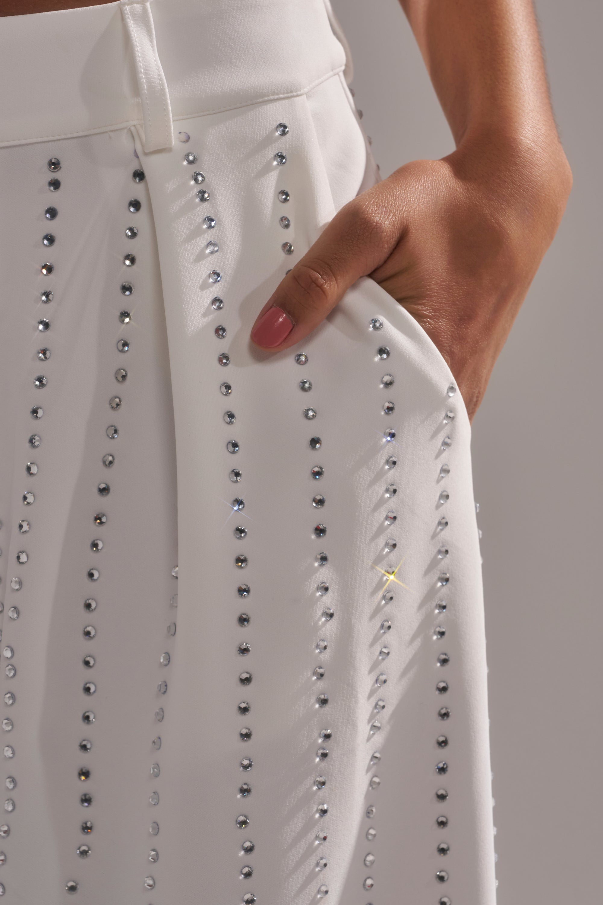 A close-up of a hand with pink nail polish resting in the pocket of the NEW YEARS QUEEN TROUSER—white, high-rise wide-leg pants adorned with vertical rows of sparkling rhinestones. 6