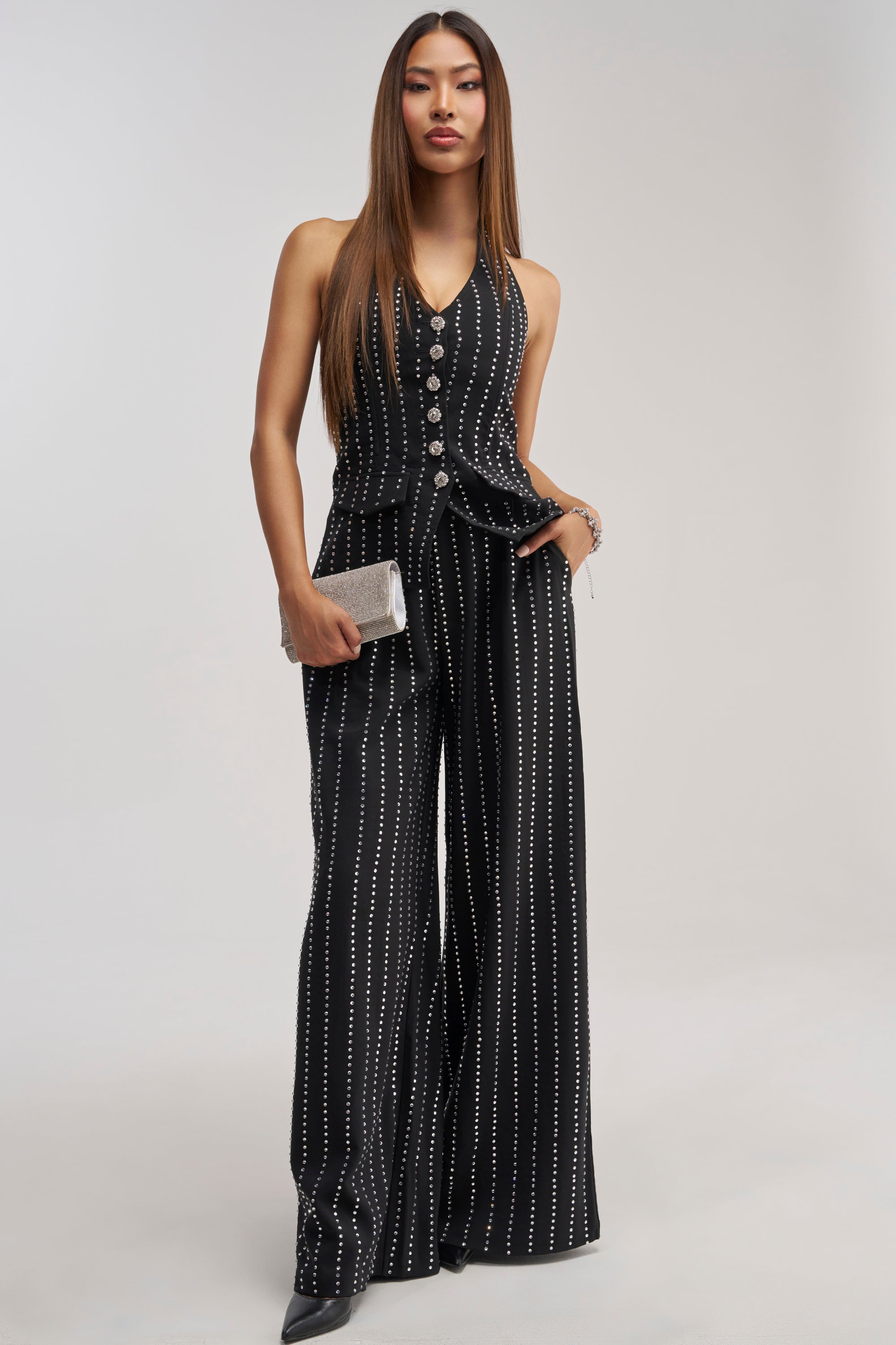 A woman in the PARTY QUEEN TROUSER—a sleeveless black jumpsuit with vertical white dotted lines and a flattering wide-leg silhouette—holds a silver clutch as she stands confidently against a plain light background, making it the perfect choice for polished formal looks.