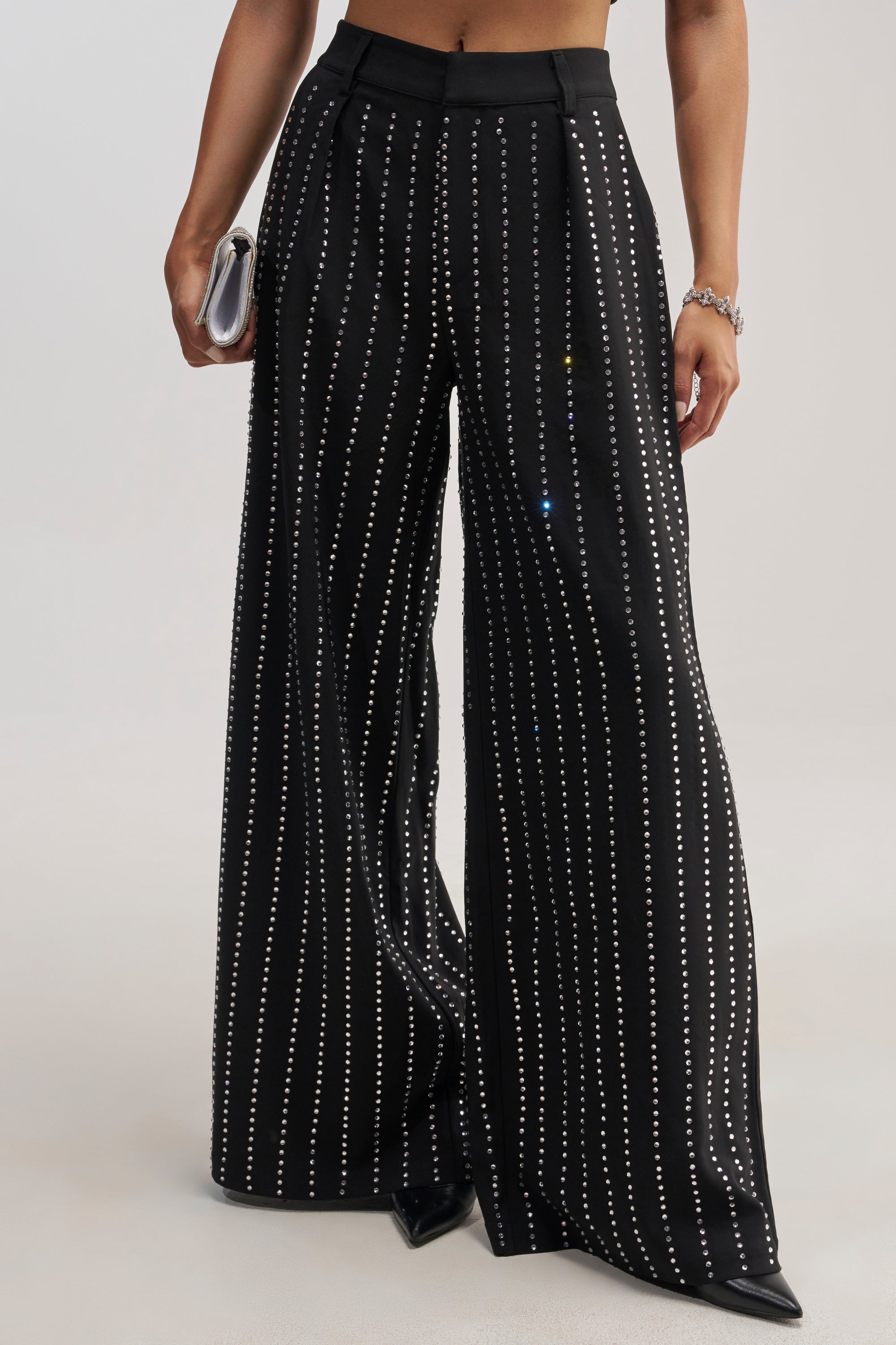 A woman wears the PARTY QUEEN TROUSER, featuring a high waist and dramatic wide-leg silhouette with vertical rows of silver studs. She holds a small silver clutch, while the formal trousers reach the floor to cover her black pointed-toe shoes. Only her torso and legs are visible.