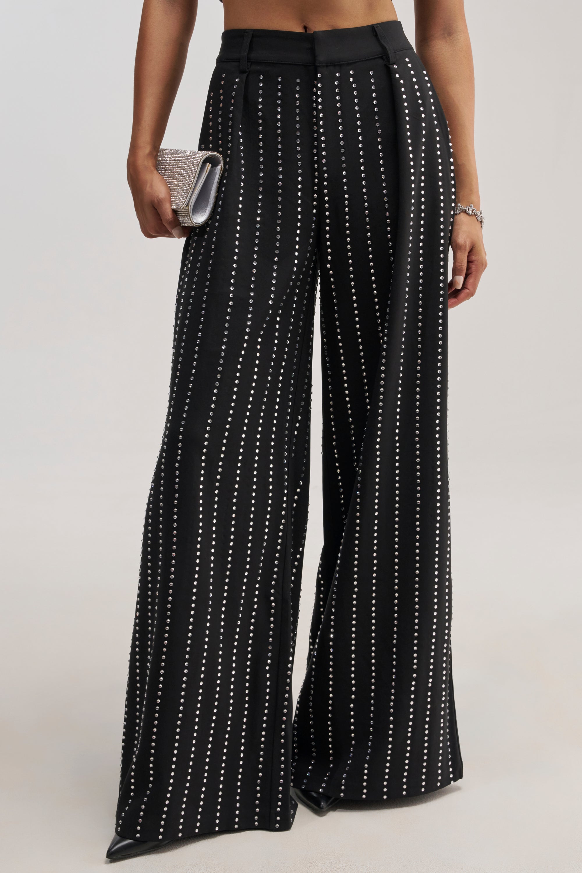 A person wearing the PARTY QUEEN TROUSER, which features a high-waisted, wide-leg silhouette in black with vertical white dotted stripes, and holding a sparkly silver clutch purse.