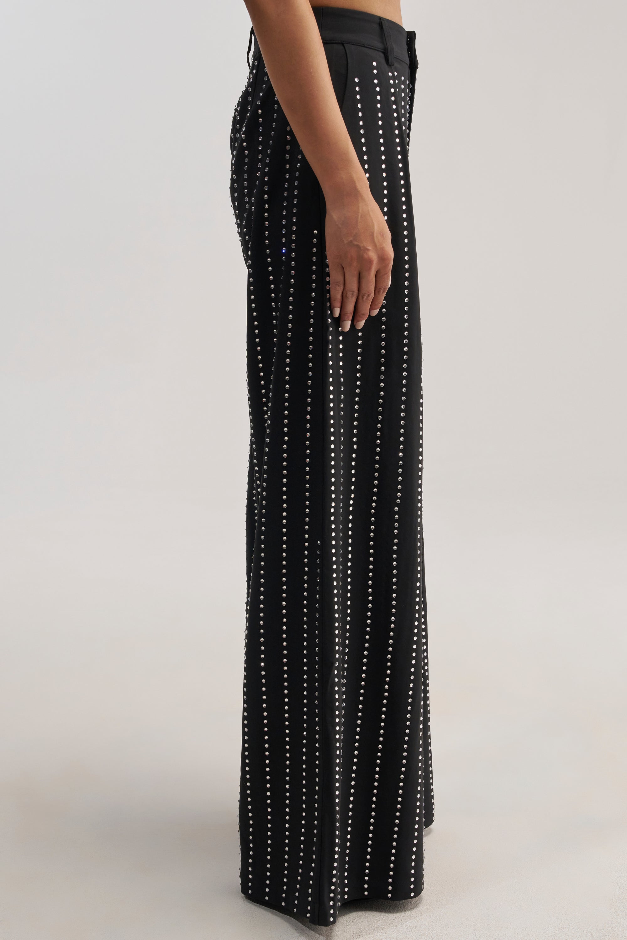 A person wearing the PARTY QUEEN TROUSER—a high-waisted, wide-leg black pant featuring vertical rows of small white dots—stands sideways against a plain light background.
