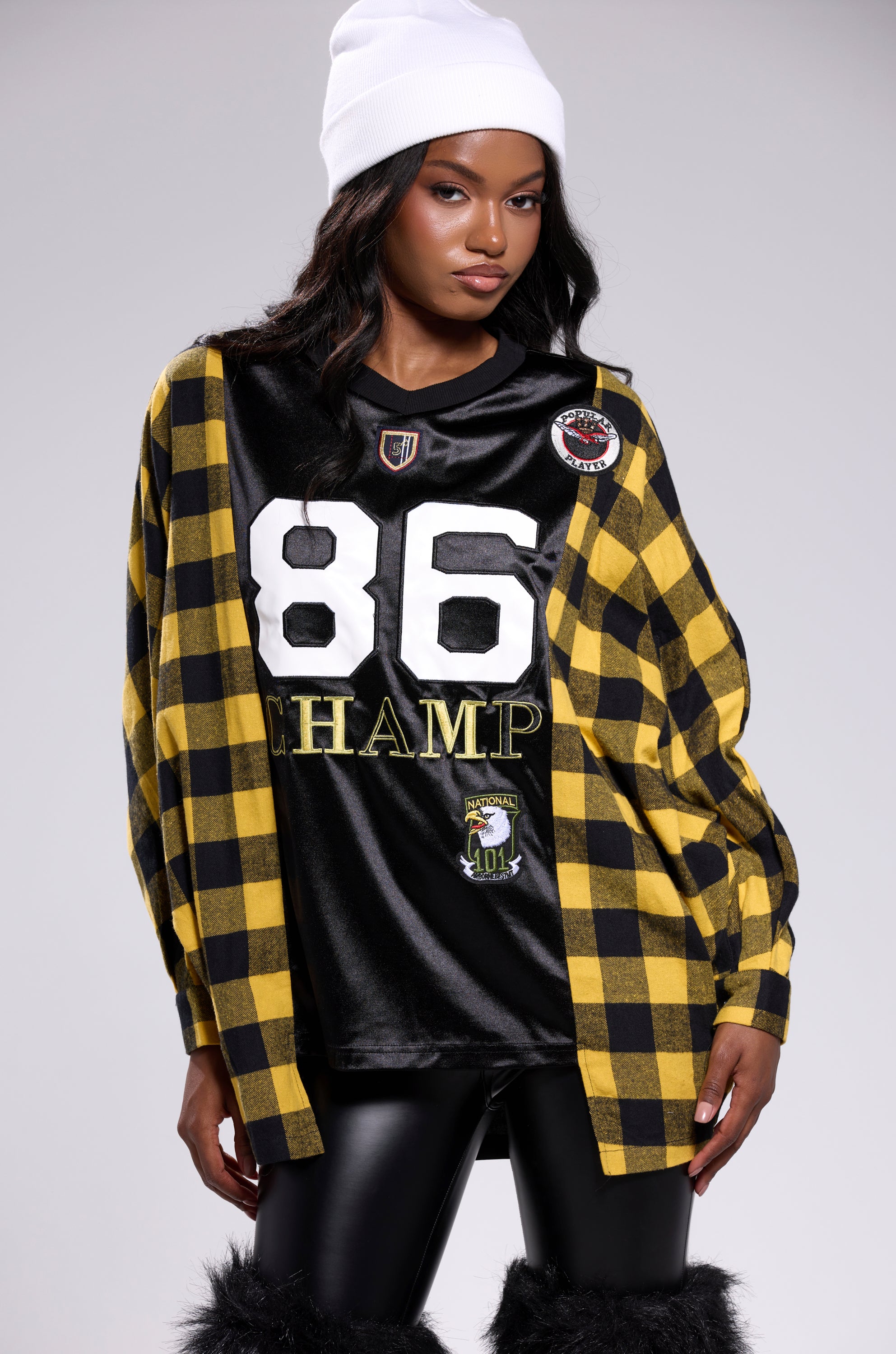 A woman in streetwear oversized style wears a white beanie, a black and yellow checkered jacket, the NEW CHAMP REBUILT JERSEY FLANNEL TOP, black leather pants, and furry boots as she poses against a plain background.