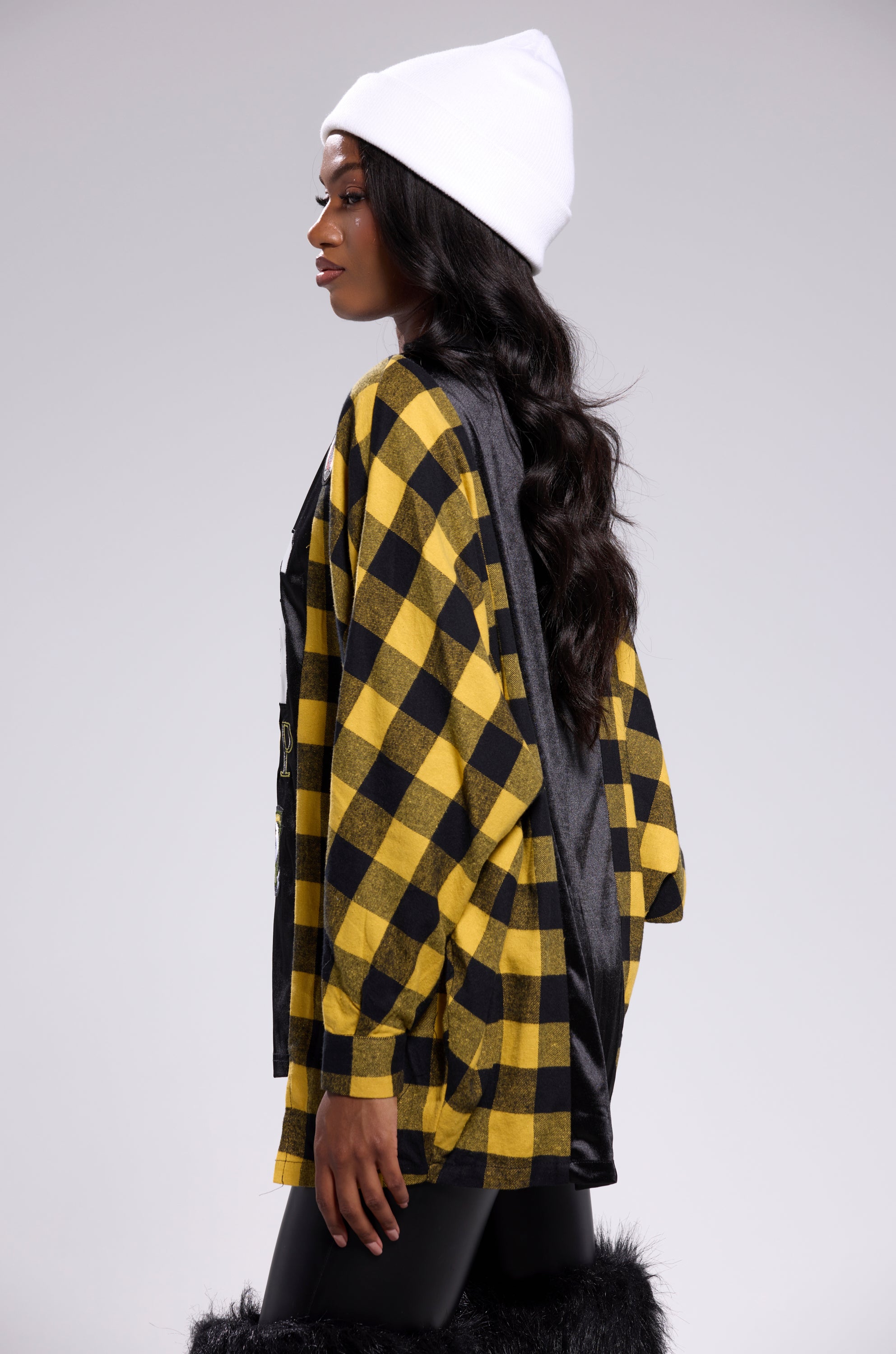 A woman in a white beanie and black leggings stands in profile against a plain background, sporting the AKIRA Label NEW CHAMP REBUILT JERSEY FLANNEL TOP and long wavy hair for an oversized streetwear look.