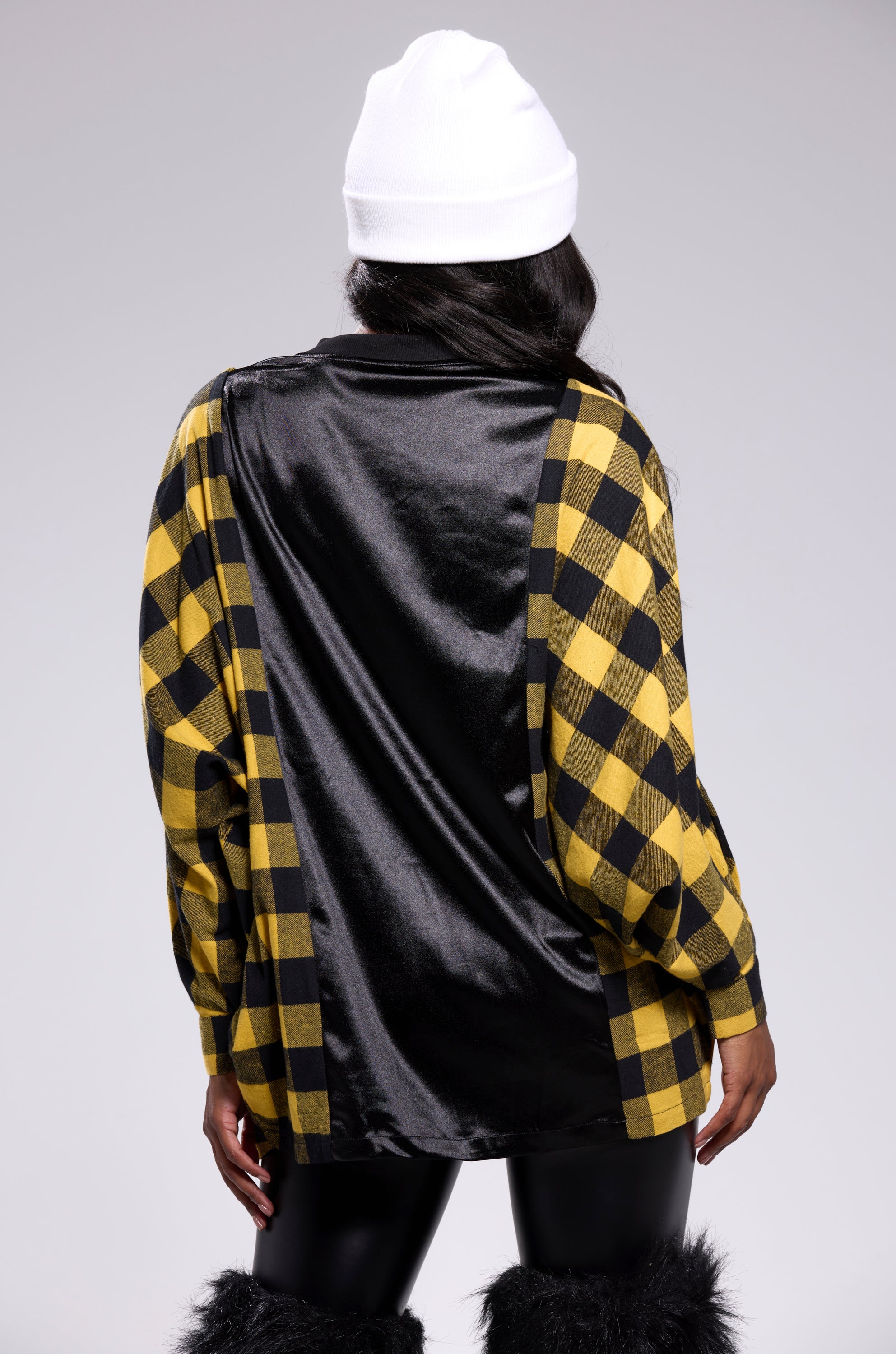 A person wearing the NEW CHAMP REBUILT JERSEY FLANNEL TOP in an oversized black and yellow checkered style, paired with a white beanie, black pants, and black furry boots is seen from behind against a plain light gray background.