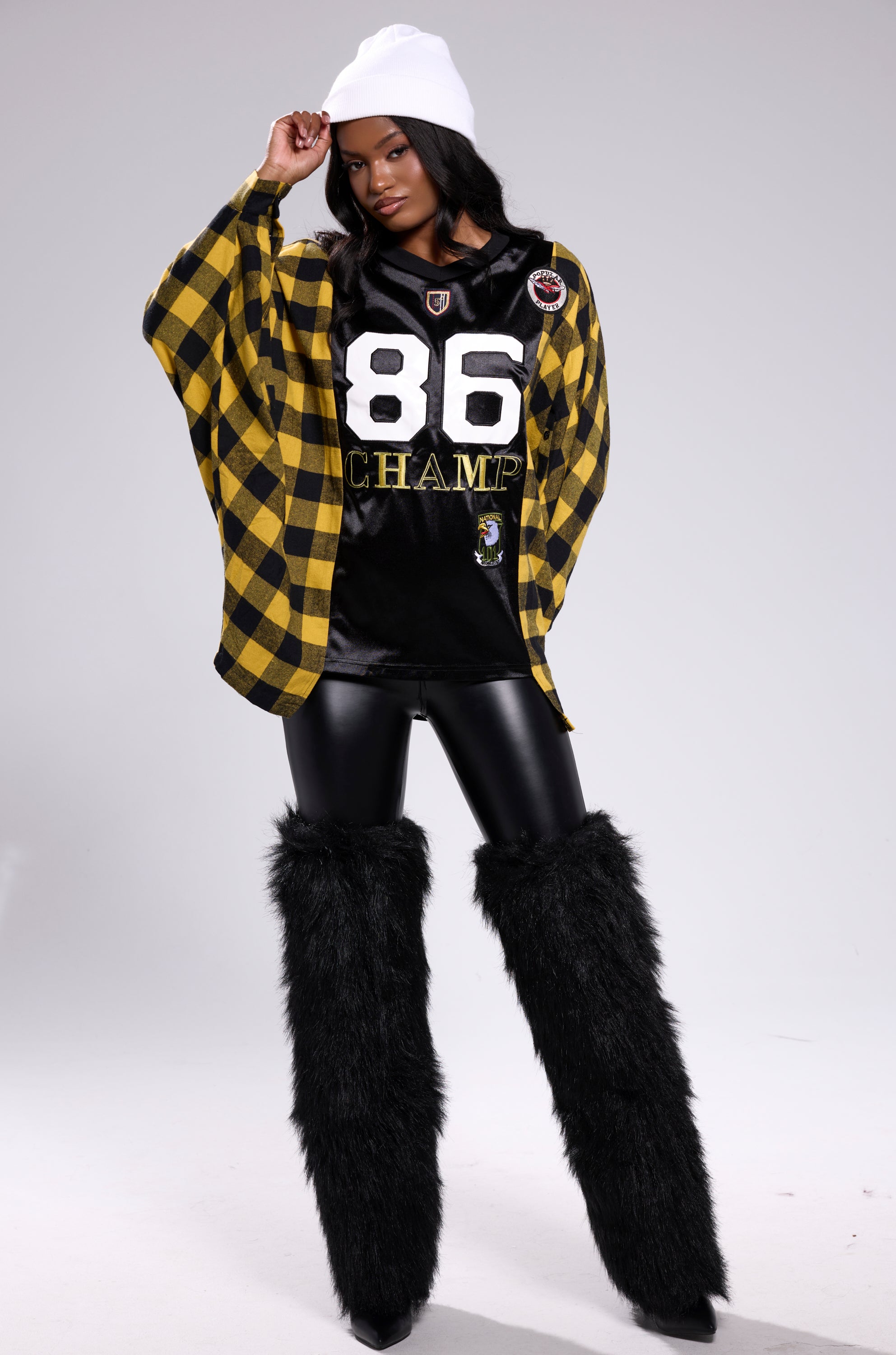 A woman poses confidently in a white beanie, streetwear oversized black and yellow checkered jacket, NEW CHAMP REBUILT JERSEY FLANNEL TOP, shiny black leggings, and tall black furry boots against a light gray backdrop.