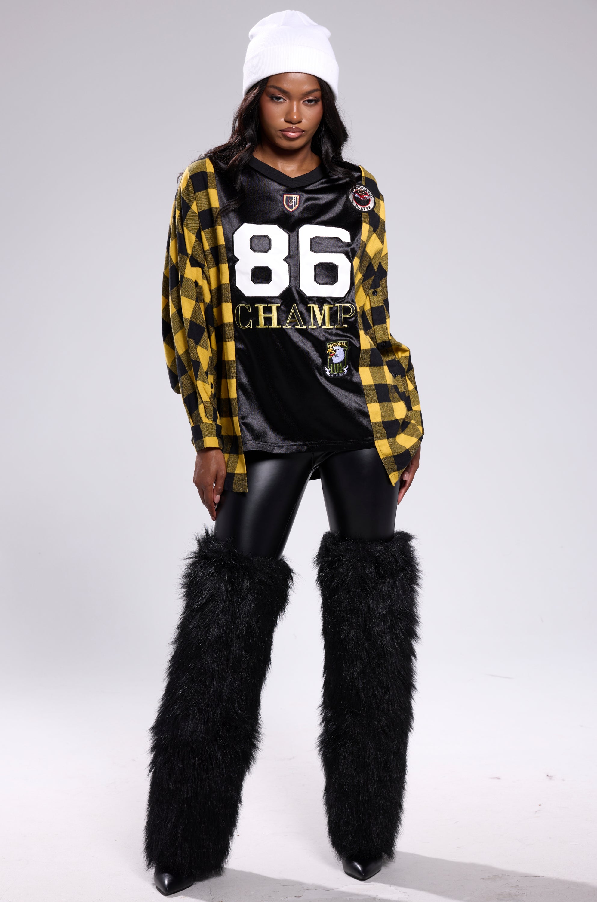 A woman poses confidently in streetwear, pairing a white beanie with the NEW CHAMP REBUILT JERSEY FLANNEL TOP over an "86 CHAMP" jersey, shiny black leggings, and tall black furry leg warmers against a plain light background.