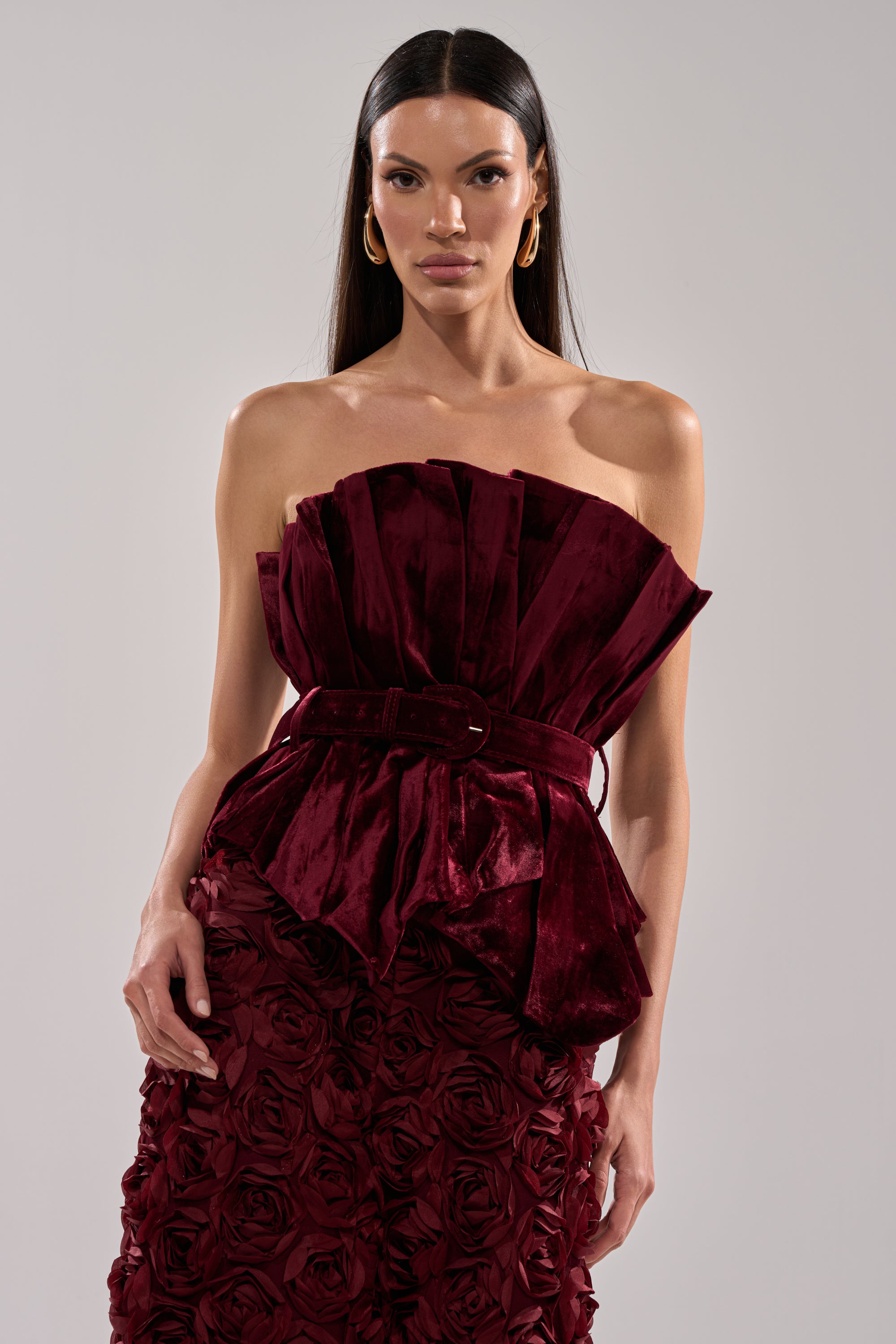 A woman wears the DRAMA DRAMA DRAMA VELVET BLOUSE, a strapless deep red velvet top with pleated sculptural details and a matching belt, paired with a textured rose-patterned skirt. She has long straight hair and gold hoop earrings.