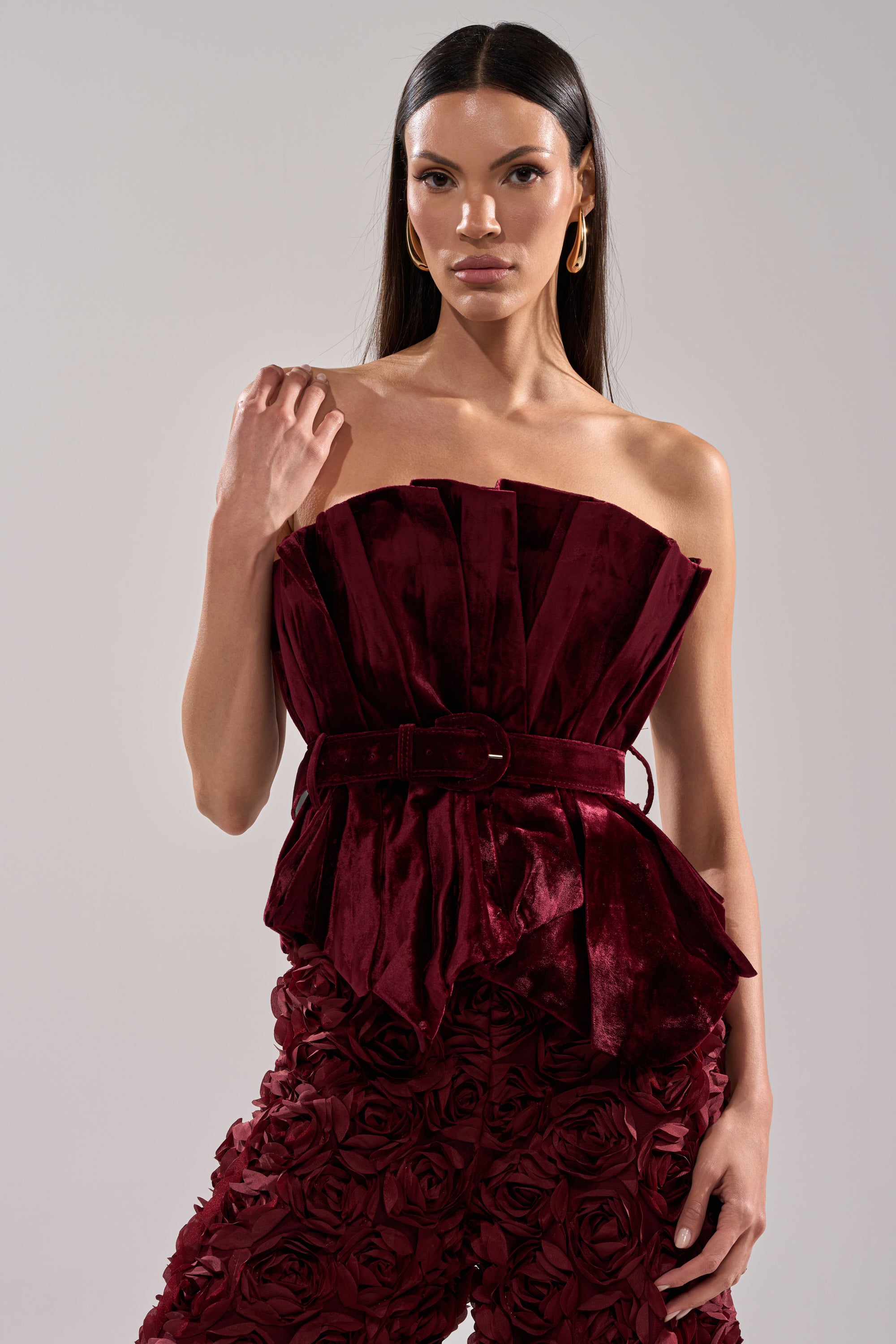 A woman with long straight hair wears the DRAMA DRAMA DRAMA VELVET BLOUSE, a strapless top with a peplum hem and belted waist, paired with matching 3D floral textured pants. She poses confidently against a plain light background.