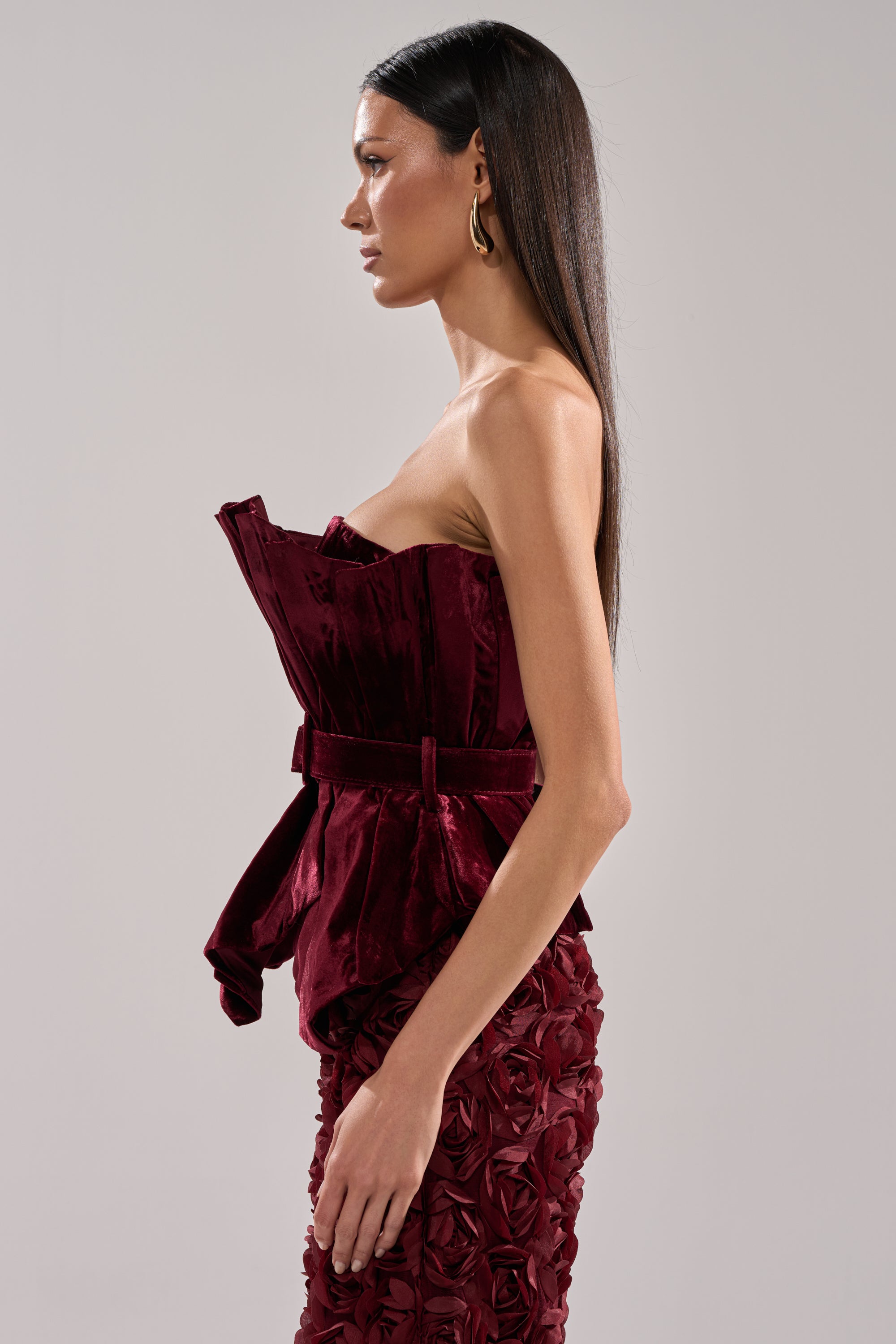 A woman stands in profile wearing the DRAMA DRAMA DRAMA VELVET BLOUSE—a burgundy strapless top with a matching belt—paired with a textured skirt featuring rose-like patterns, styled with long straight hair and gold hoop earrings against a plain backdrop.
