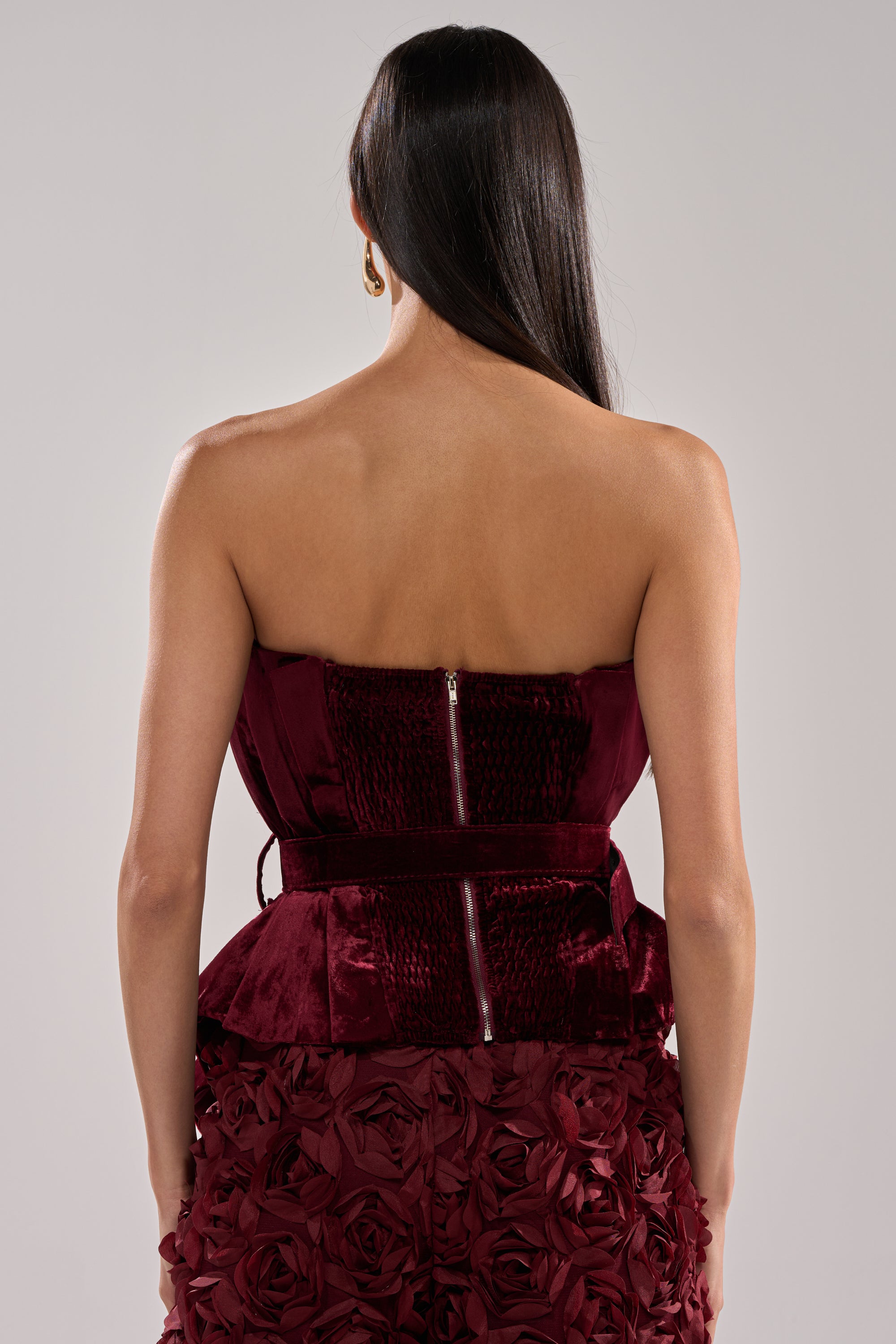 A woman with long dark hair is seen from behind, wearing the DRAMA DRAMA DRAMA VELVET BLOUSE—a strapless deep red velvet top with a zipper detail on the bodice and a textured floral skirt.