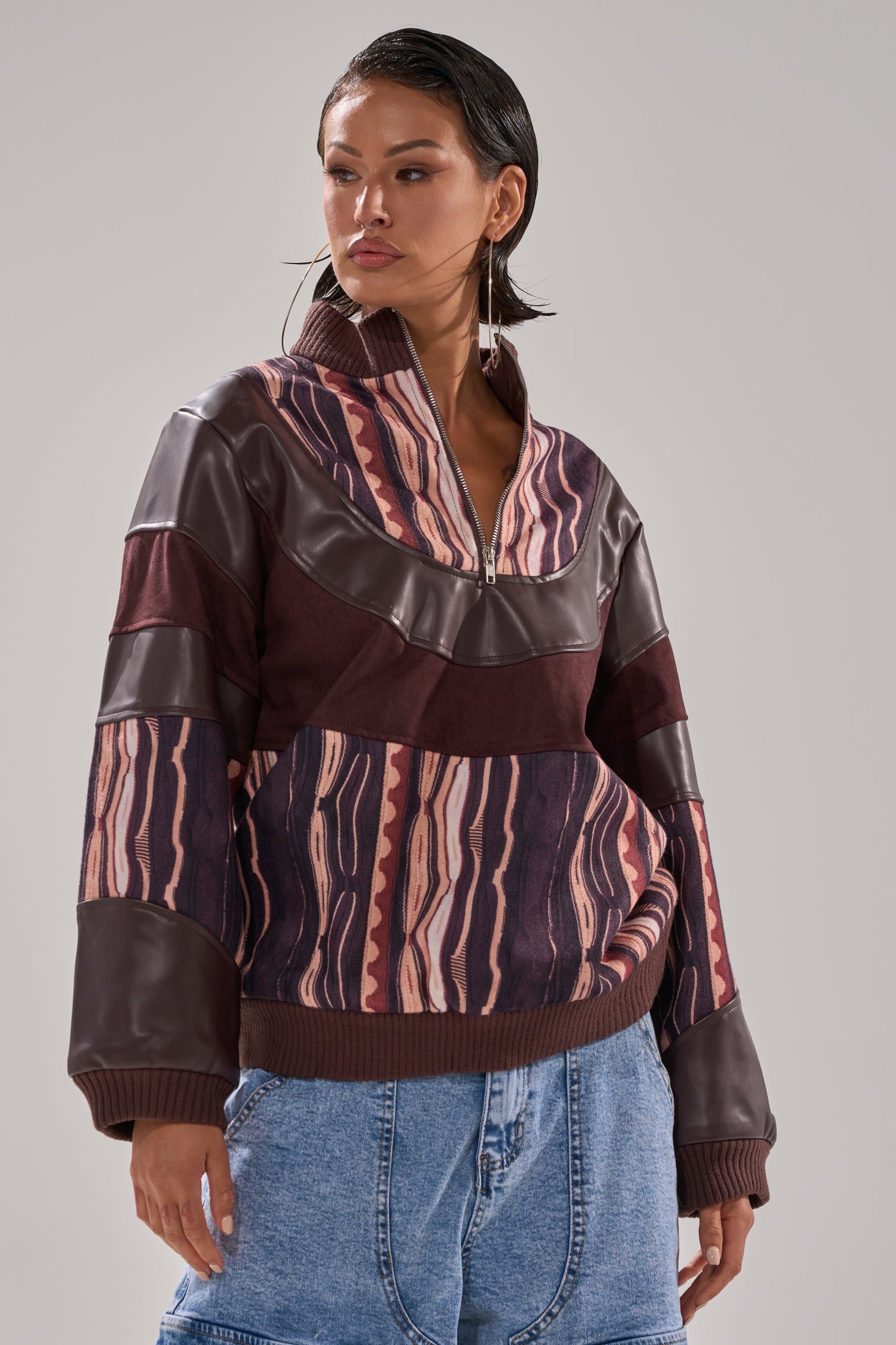 A woman wears the CRAZY IN LOVE FAUX LEATHER SWEATSHIRT, featuring a half-zip and abstract striped print in burgundy and pink, paired with light blue jeans against a plain gray background.