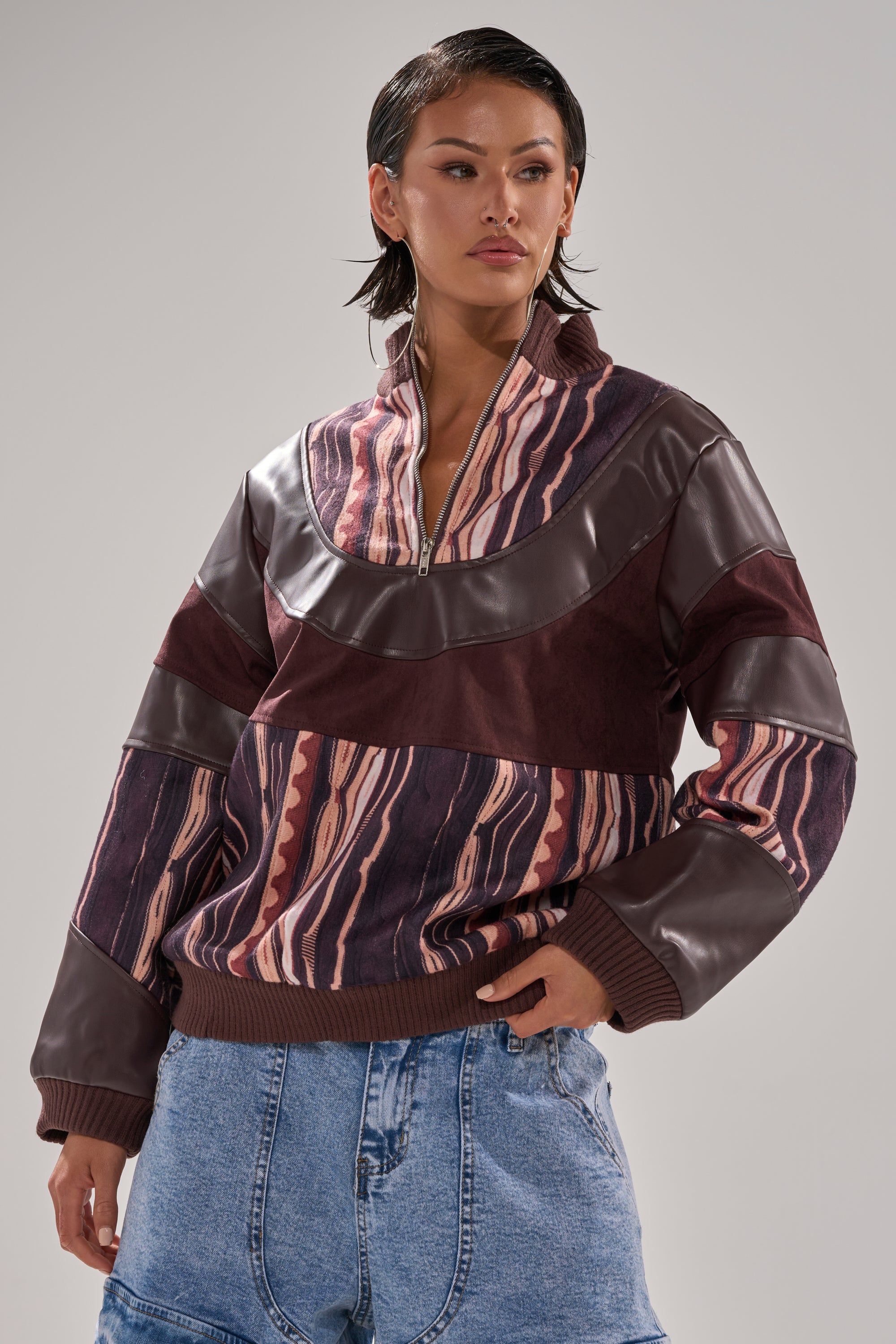 A person with short dark hair wears the CRAZY IN LOVE FAUX LEATHER SWEATSHIRT, featuring an abstract striped print in maroon, brown, and pink with faux leather details, paired with blue denim jeans against a plain light background.