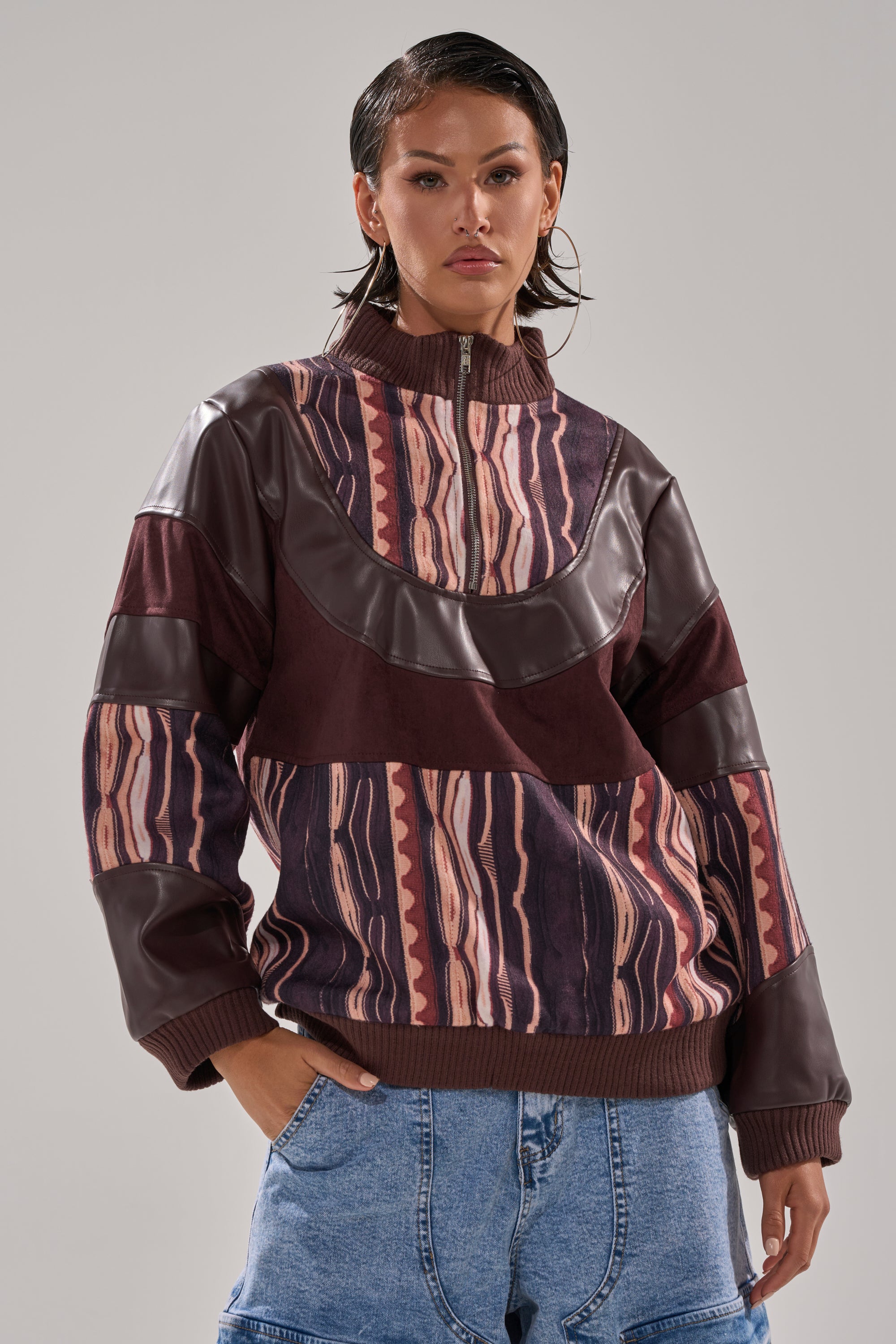 A person with wet-look hair wears the CRAZY IN LOVE FAUX LEATHER SWEATSHIRT—a maroon and brown zip-up jacket with faux leather accents—and wide blue denim jeans, posing confidently against a neutral background.