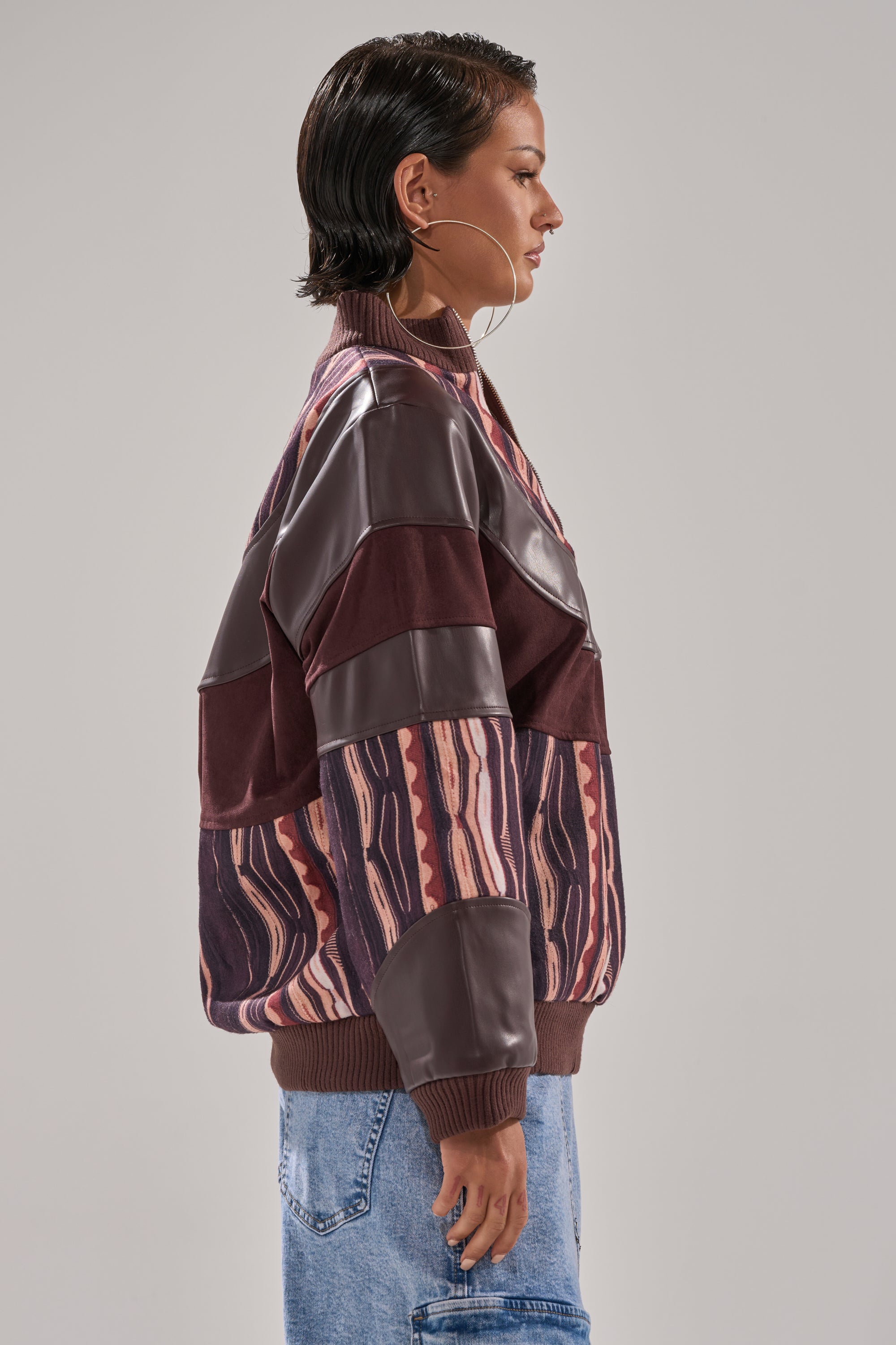 A woman with short dark hair stands sideways against a plain background, wearing the CRAZY IN LOVE FAUX LEATHER SWEATSHIRT with abstract striped print, paired with light blue jeans and large hoop earrings.