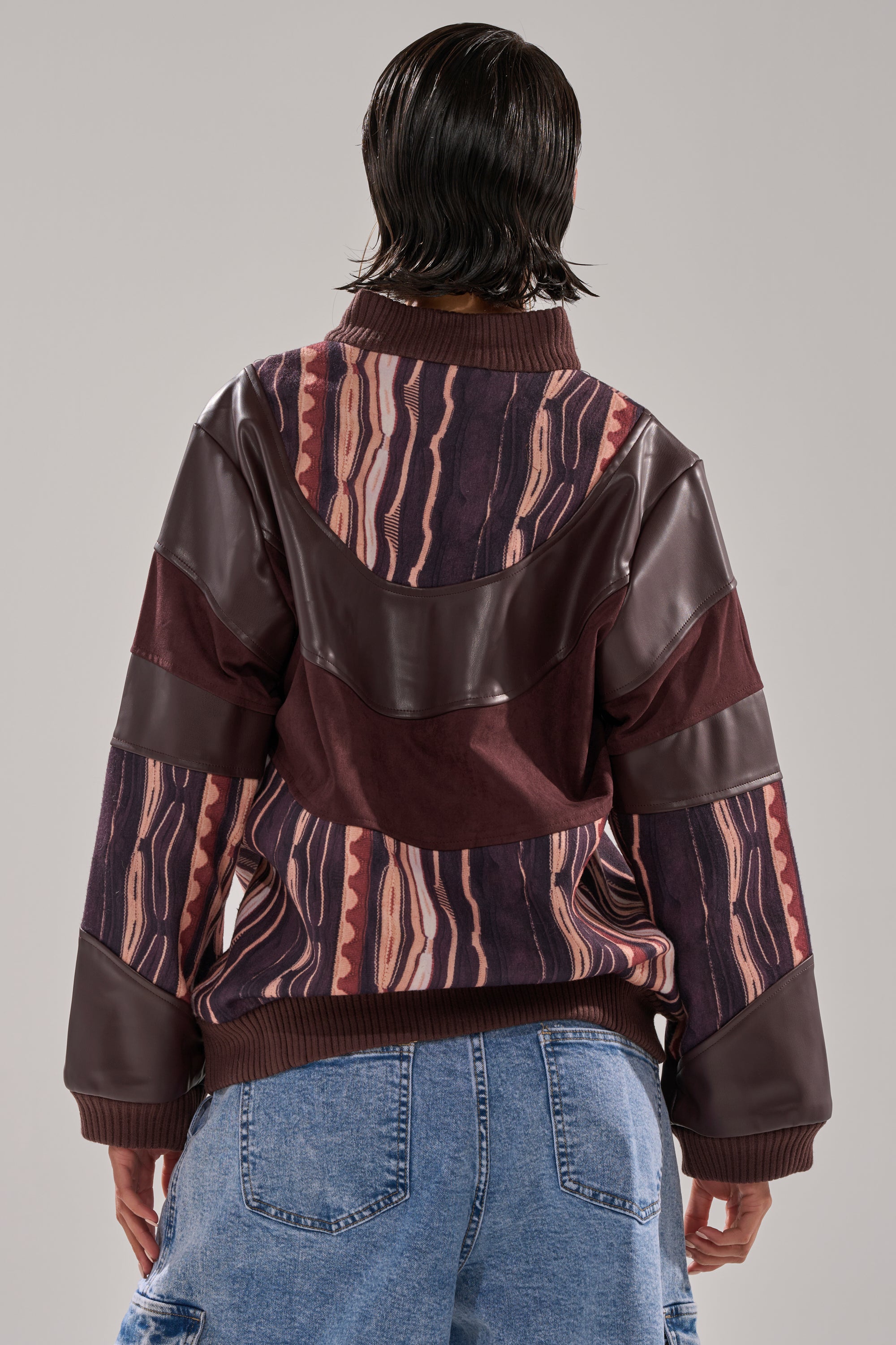 A person with short, wet-look dark hair is shown from the back, wearing blue jeans and the CRAZY IN LOVE FAUX LEATHER SWEATSHIRT—patterned in maroon and brown faux leather panels—against a plain light background.