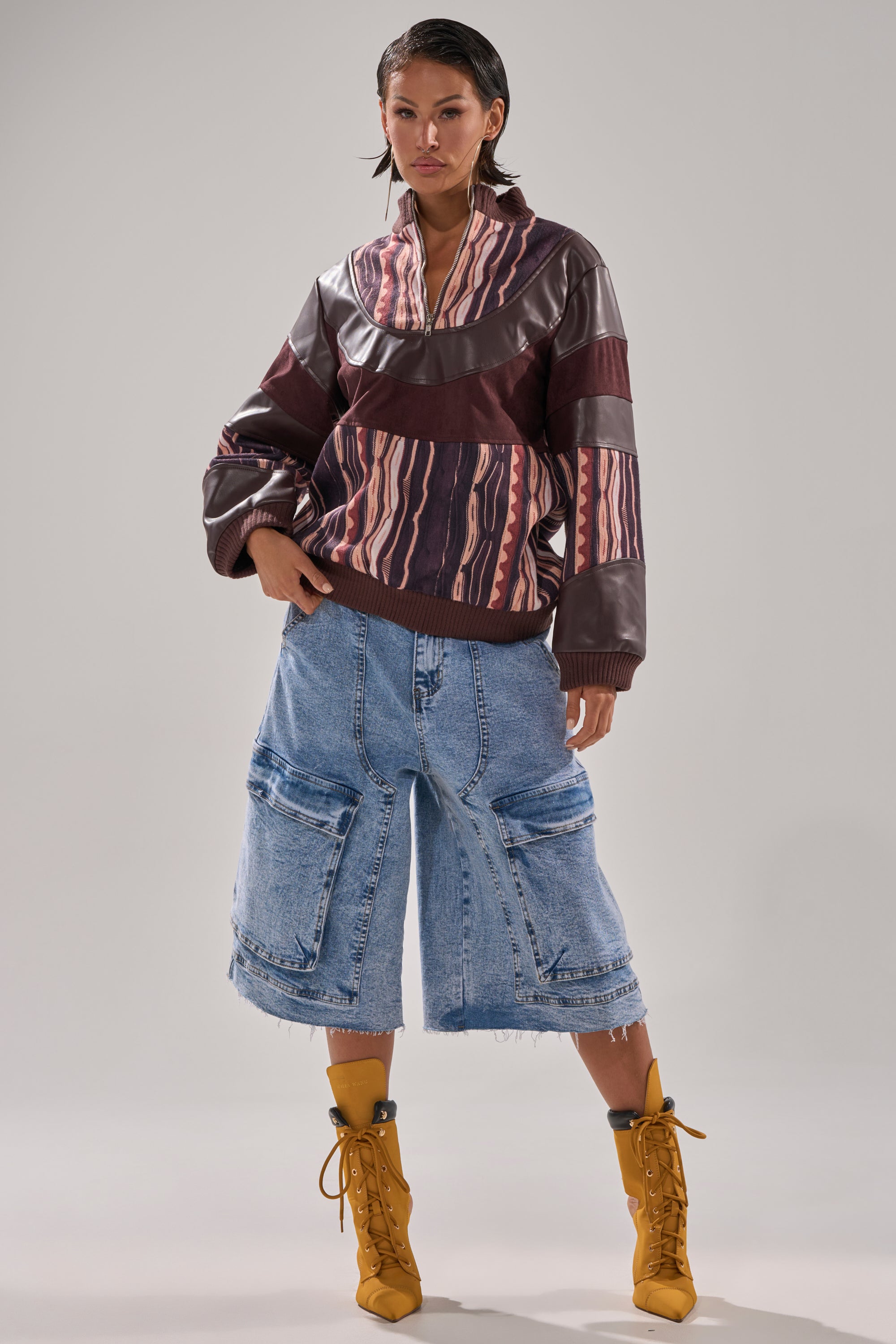A person in the CRAZY IN LOVE FAUX LEATHER SWEATSHIRT, loose knee-length denim shorts with large pockets, and mustard yellow lace-up boots stands confidently against a plain background.