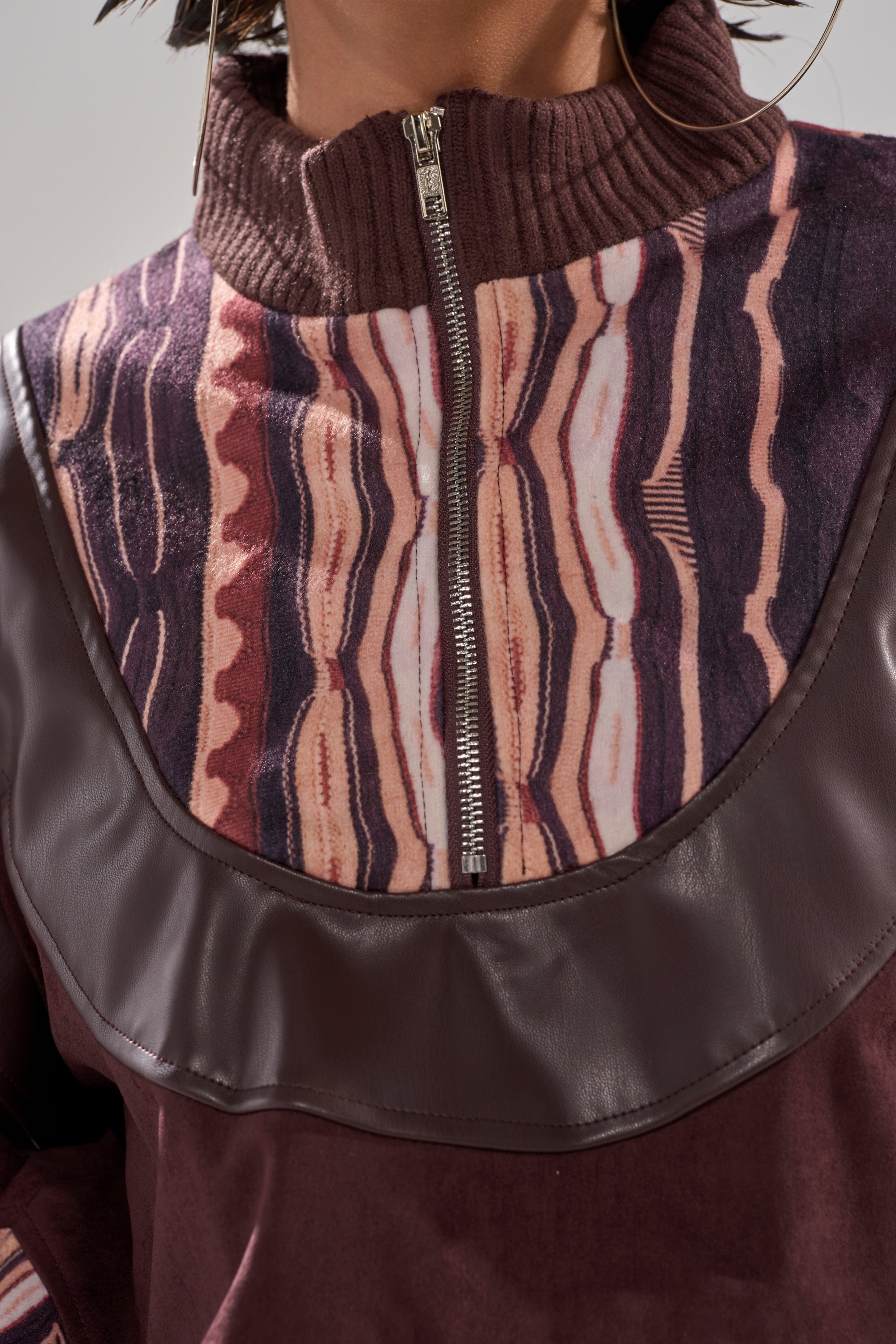 A close-up of a woman's torso wearing the CRAZY IN LOVE FAUX LEATHER SWEATSHIRT, featuring a relaxed fit, high collar with zipper, maroon and pink wavy abstract stripes, and large hoop earrings.