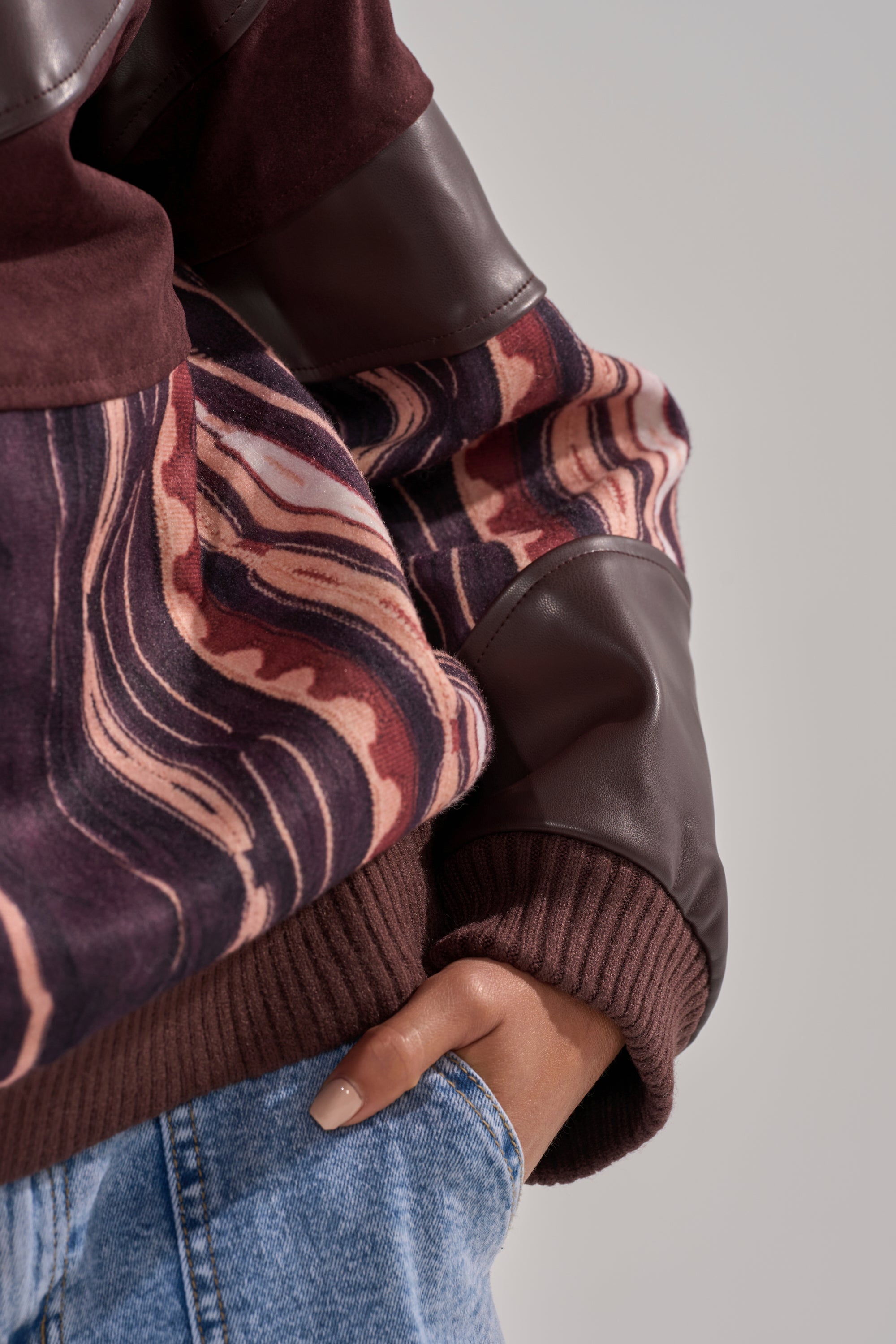 Wearing the CRAZY IN LOVE FAUX LEATHER SWEATSHIRT in maroon and brown with abstract pink, brown, and cream striped sleeves, paired with blue jeans and hand in pocket.