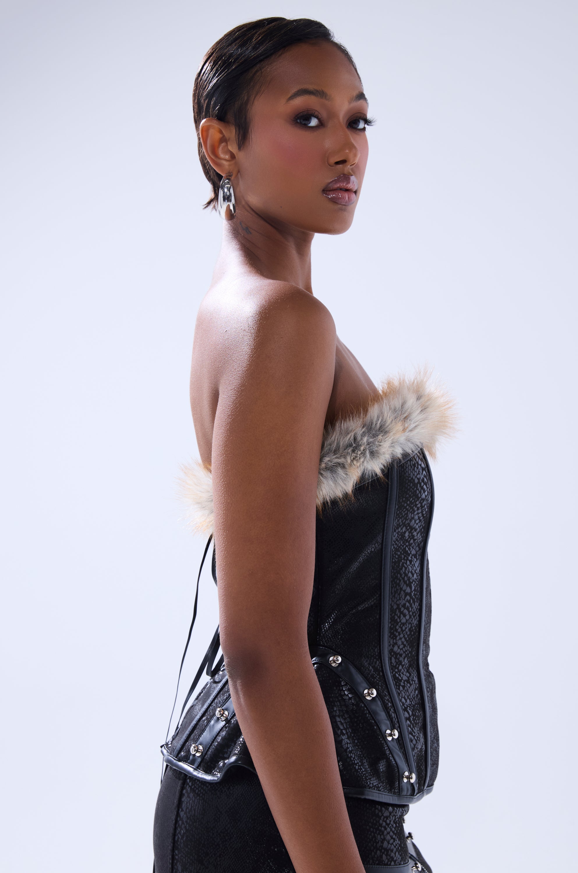 A woman poses confidently in the TELL ME WHAT YOU WANT CORSET WITH FUR—black faux leather with fur trim and metallic studs. With short, slicked-back hair and hoop earrings, she looks over her shoulder against a light background.