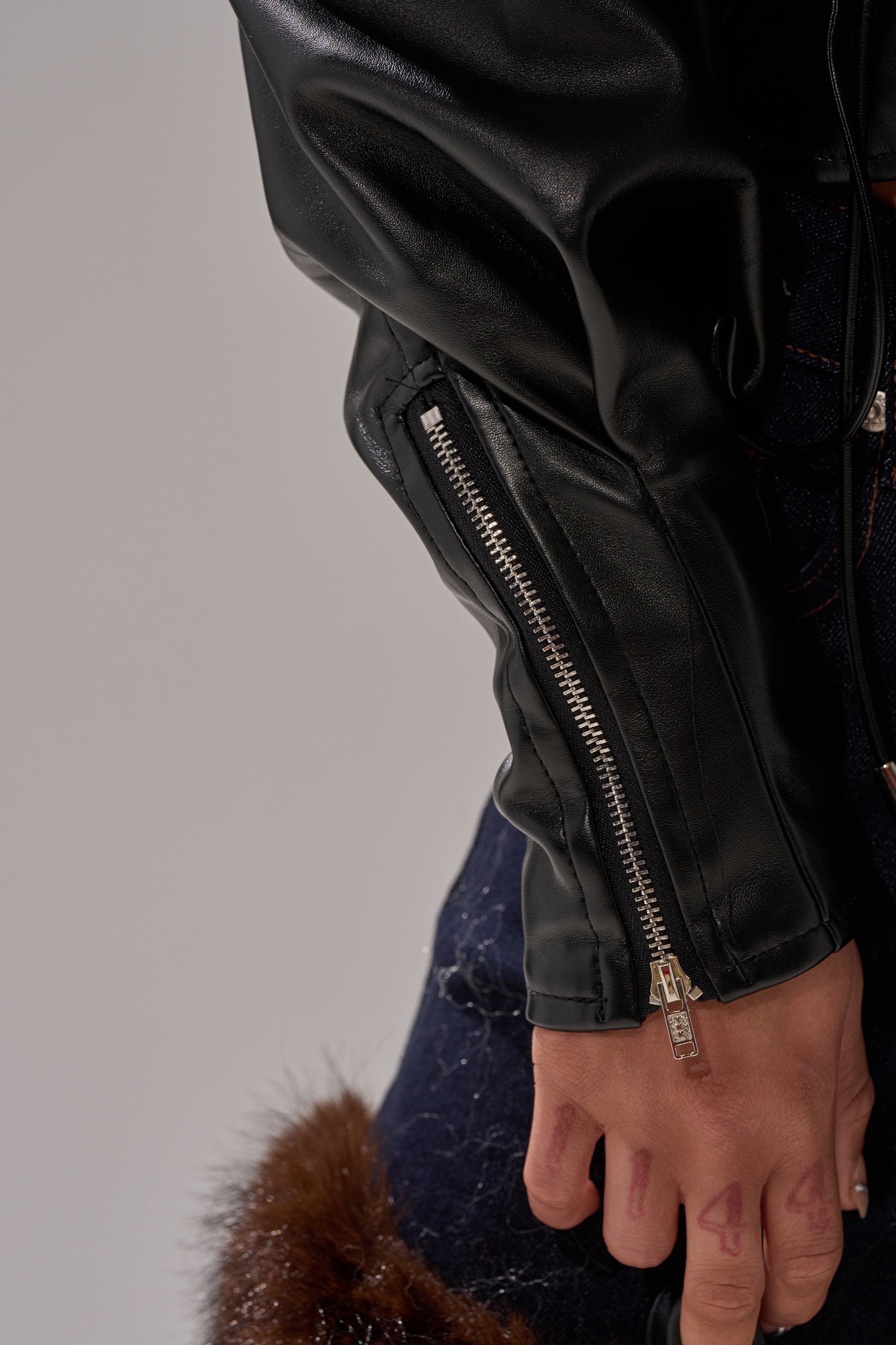 A close-up of a hand with red ink numbers near the knee, wearing the AUDREY FAUX LEATHER BLOUSE featuring a visible zipper, paired with dark jeans detailed with brown fur trim.