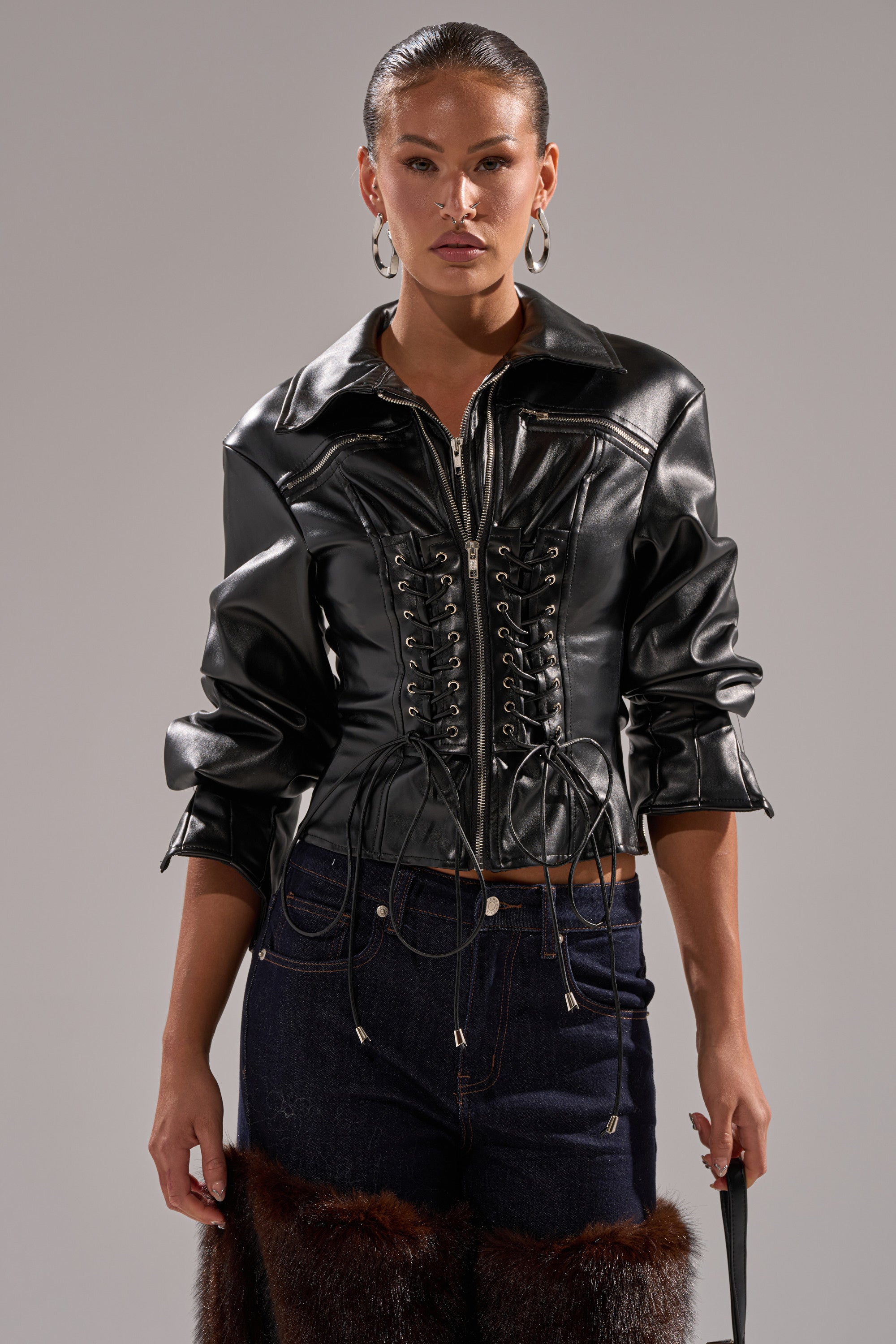 A woman models the AUDREY FAUX LEATHER BLOUSE in shiny black, styled with dark blue jeans with fur-trimmed thighs, against a plain gray background.