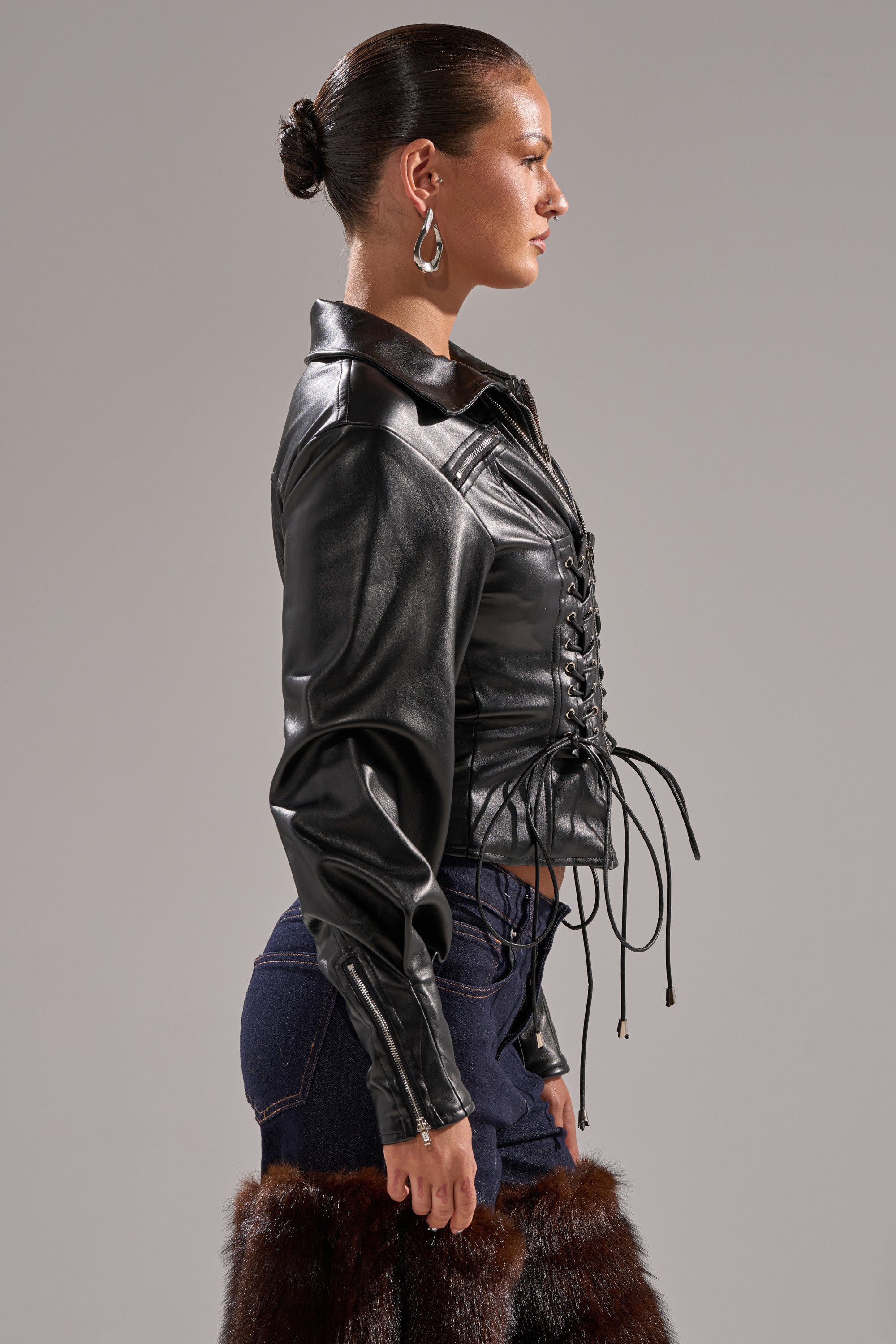 A person with slicked-back hair, silver earrings, dark blue jeans, brown fur-trimmed boots, and the AUDREY FAUX LEATHER BLOUSE stands in profile against a gray background.